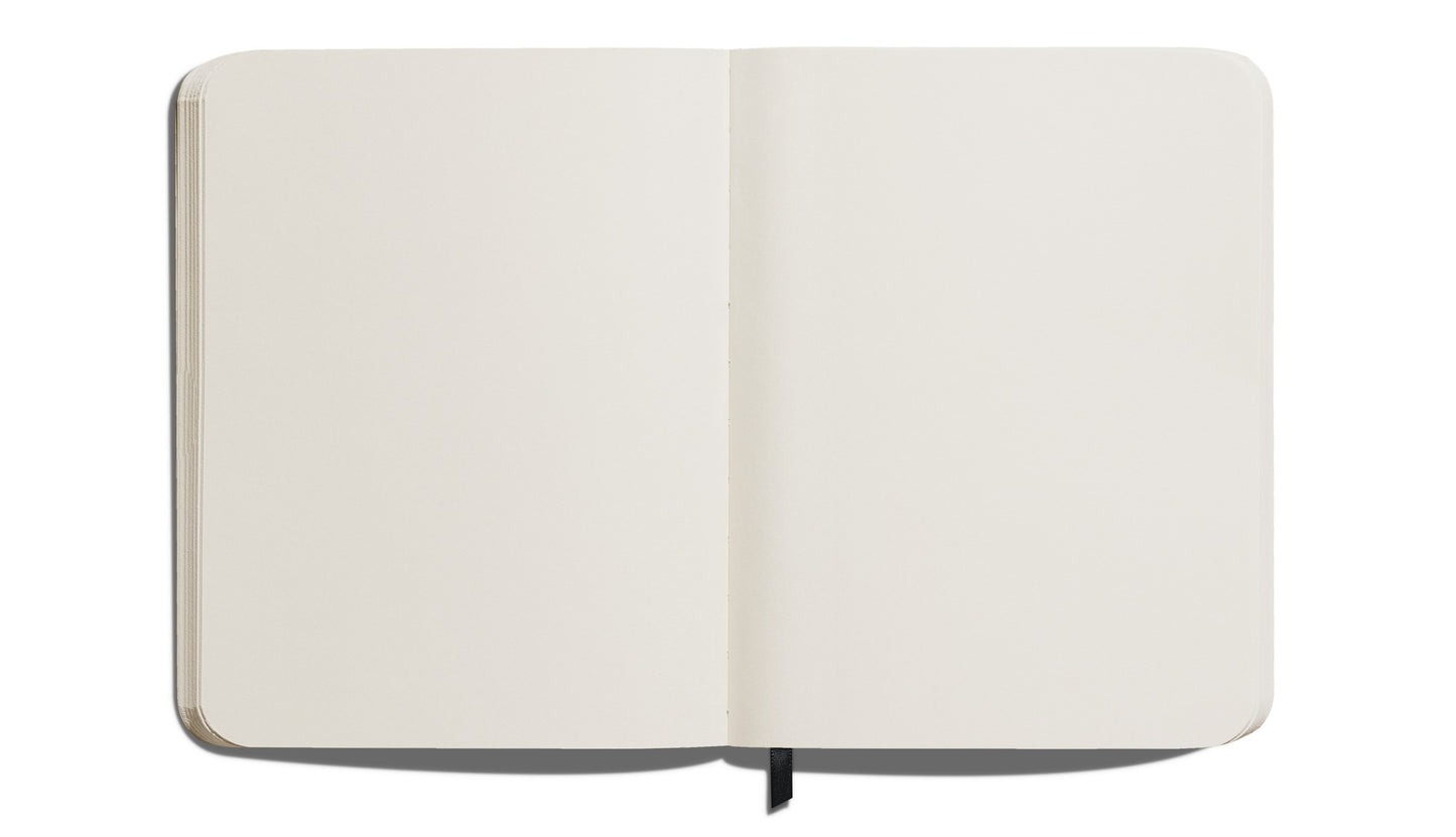 A Shinola Large Hard Plain Journal in black is shown open from above, displaying its blank cream, acid-free pages. A black ribbon bookmark appears at the bottom center.