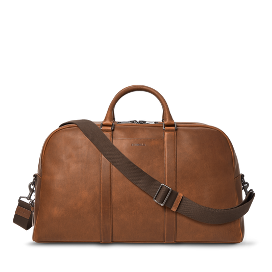 The Shinola Runwell Duffle Bag in Medium Brown is crafted from premium leather, with sturdy handles and an adjustable, detachable shoulder strap. Displayed on a white background, it's an ideal spacious carry-on for any journey.