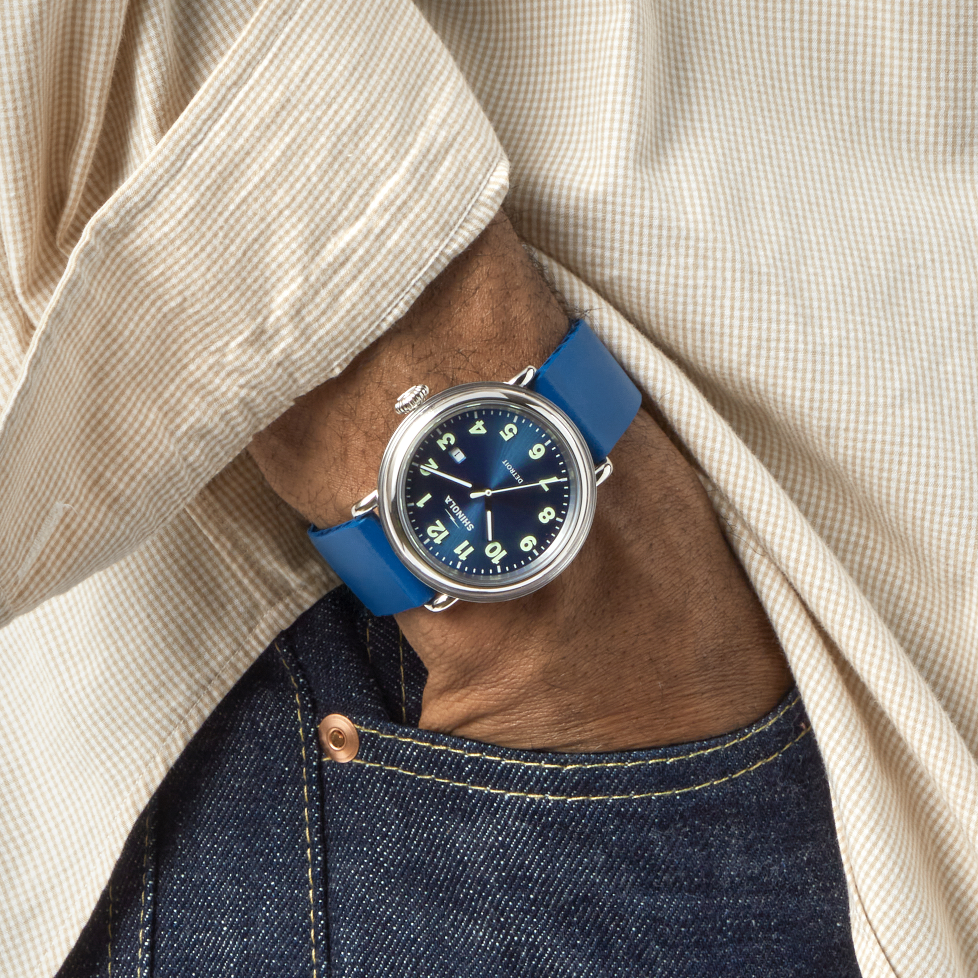 A person in a beige checkered shirt and blue jeans has their hand in their pocket, showcasing the Shinola Detrola Watch - Denim Blue with its dark face.