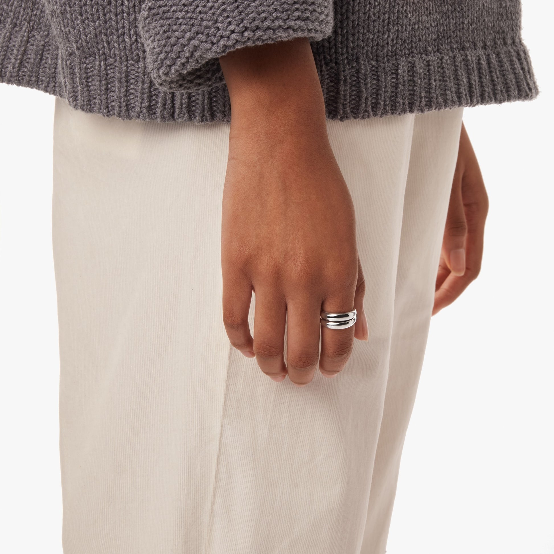 A person in a gray knit sweater and light pants is shown from the waist down, wearing the Shinola Signature Link Adjustable Ring - Sterling Silver, a bold statement piece, on their middle finger.