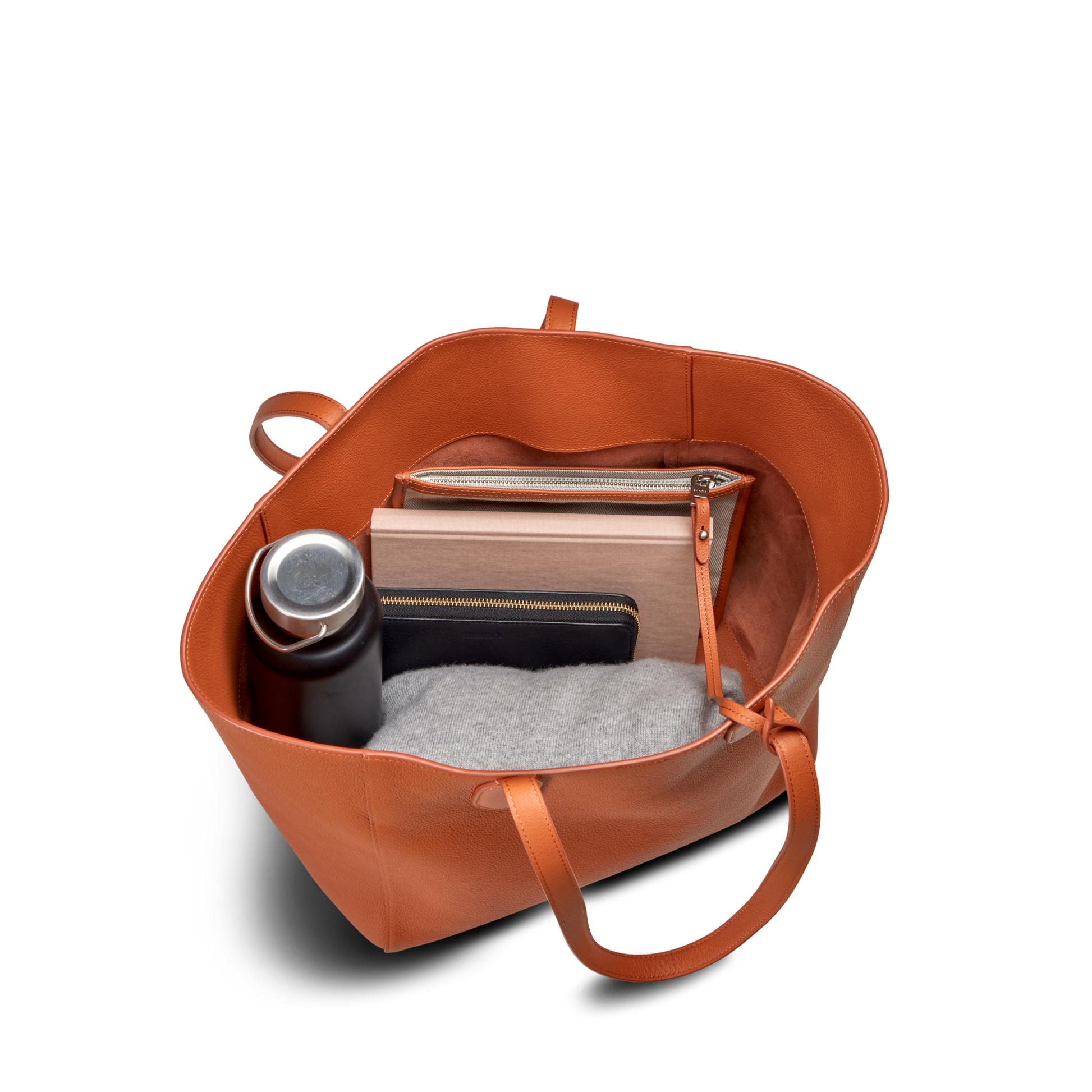The Shinola Runabout Tote Bag in Rust, shown open from above on a white background, fits daily essentials like a water bottle, grey sweater, tablet or laptop, and two zipper pouches—stylish and lightweight for everyday use.