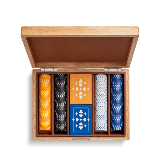 The Shinola Lacquered Wood Poker Set in Cream/Blue features a leather-lined case with an open lid, custom poker chips in yellow, black, white, and blue, plus two decks of geometric-patterned cards—one orange and one blue.