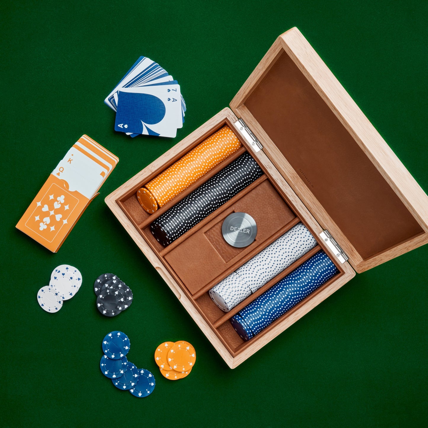 The Shinola Lacquered Wood Poker Set in Cream/Blue rests open on a green surface, displaying custom poker chips in four colors, a dealer button, and cards. Separate card stacks and scattered chips sit beside the elegant case.