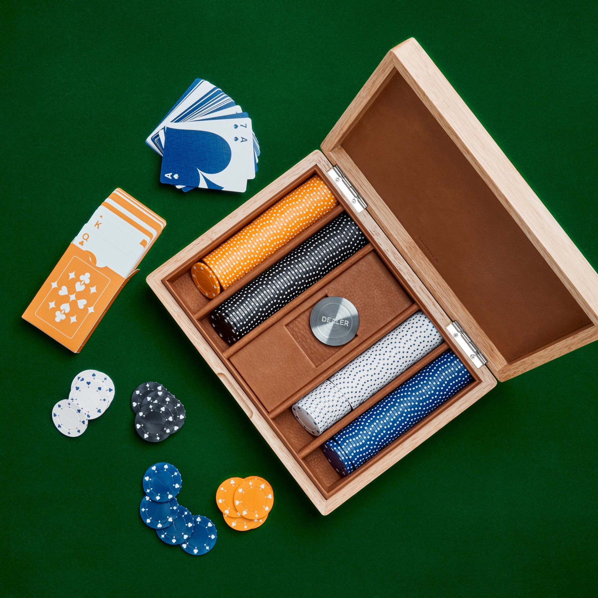 The Shinola Lacquered Wood Poker Set in Cream/Blue rests open on a green surface, displaying custom poker chips in four colors, a dealer button, and cards. Separate card stacks and scattered chips sit beside the elegant case.
