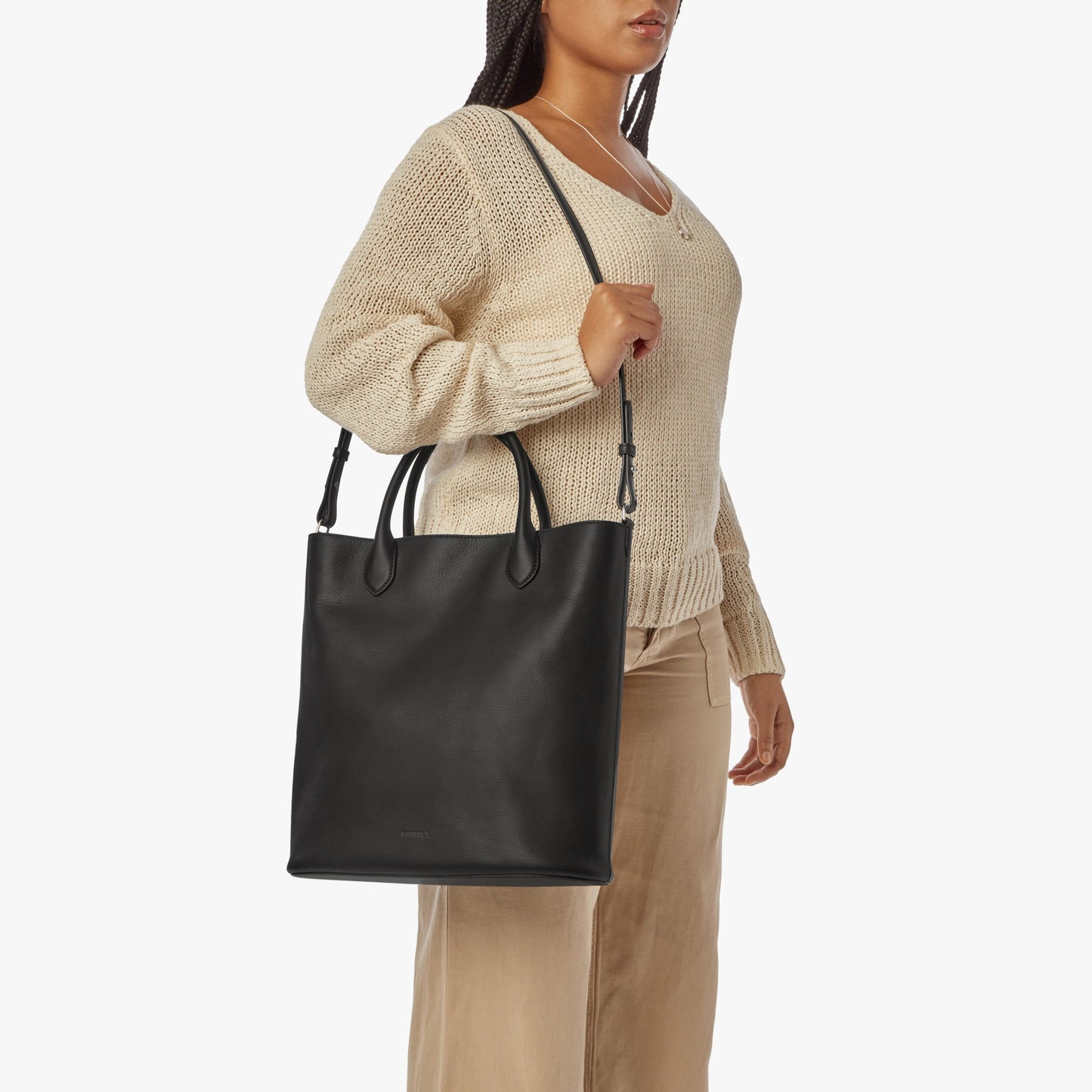 A woman wearing a beige knit sweater and tan pants carries the Shinola 12 Hour Tote Bag - Black, an everyday leather tote with rolled handles and a detachable shoulder strap, over her shoulder.