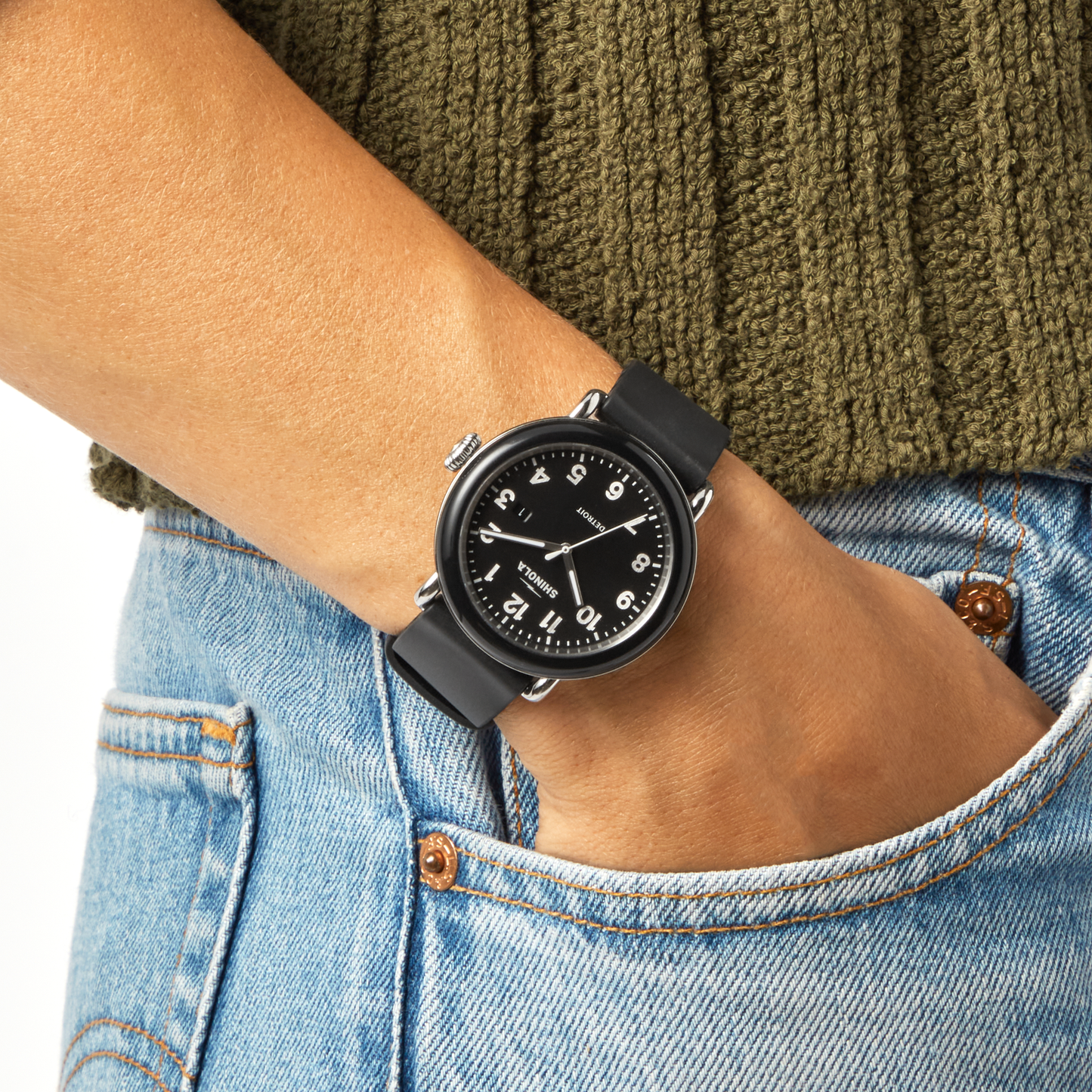 A Shinola Detrola Watch - Black with a matching band and face rests on someone's wrist, paired with a green knitted sweater and blue jeans, highlighting classic American design.
