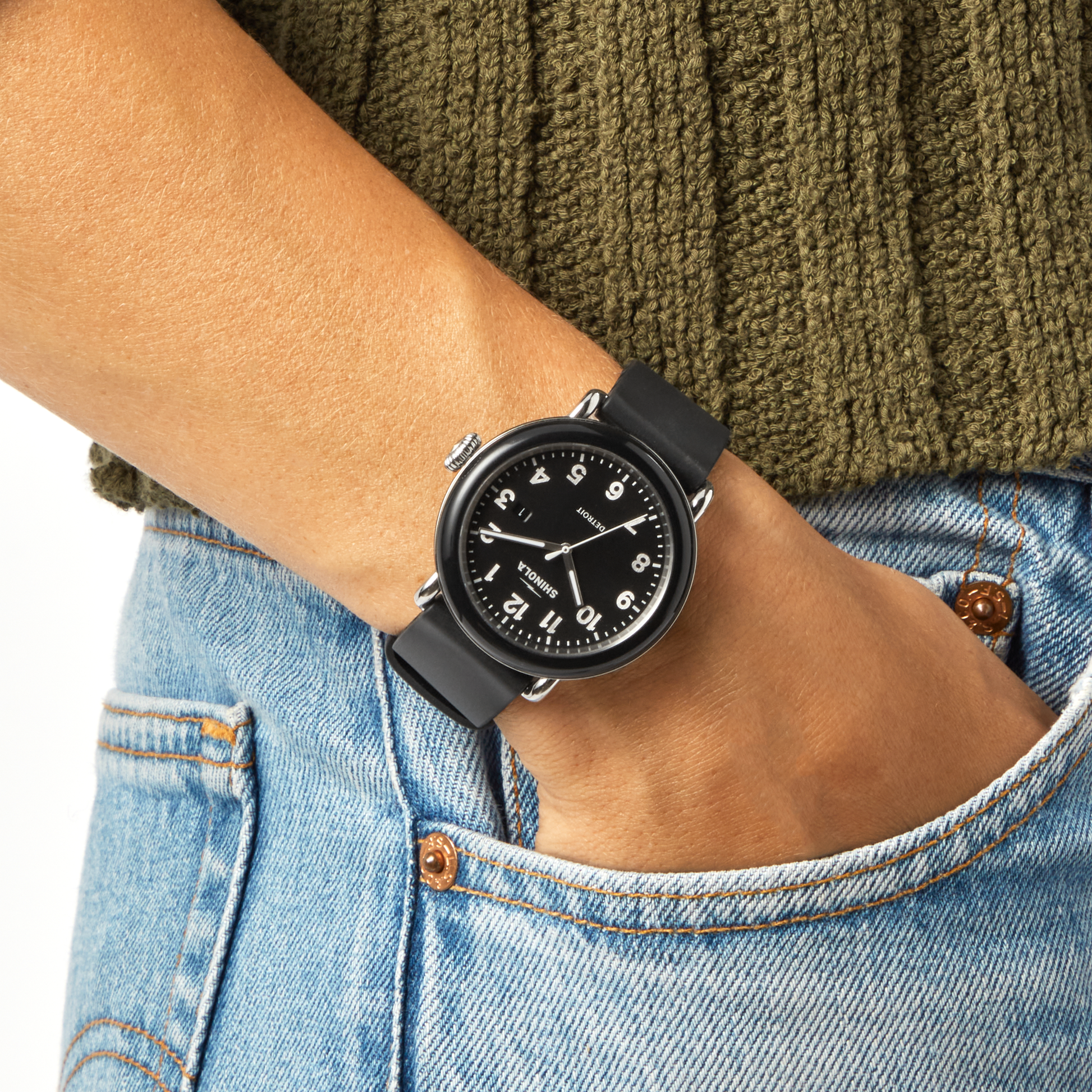 A Shinola Detrola Watch - Black with a matching band and face rests on someone's wrist, paired with a green knitted sweater and blue jeans, highlighting classic American design.
