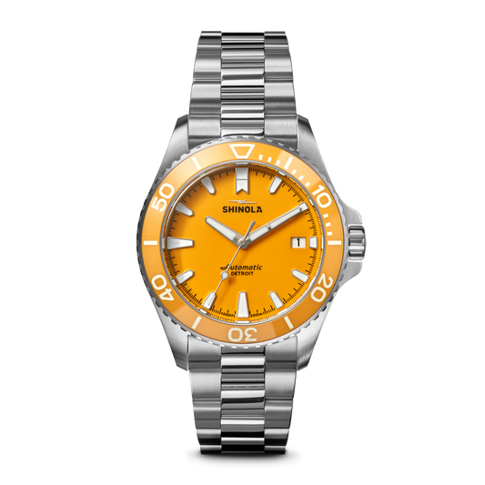 The Shinola Lake Huron Monster Automatic Dive Watch features a stainless steel case, silver metal band, bright orange face, white hour markers and hands, minute indices, and the Shinola name above the center.