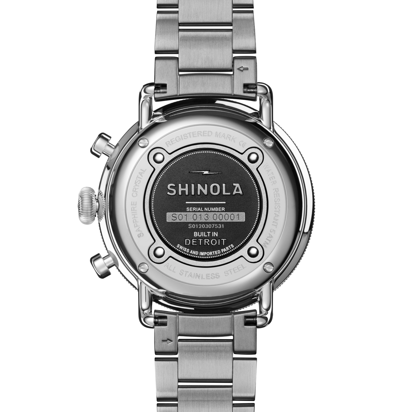 The back of the Shinola Canfield Chronograph Watch - Dark Olive displays the engraved brand name, serial number, and “Built in Detroit” text on its stainless steel case, along with the visible metal bracelet and clasp.