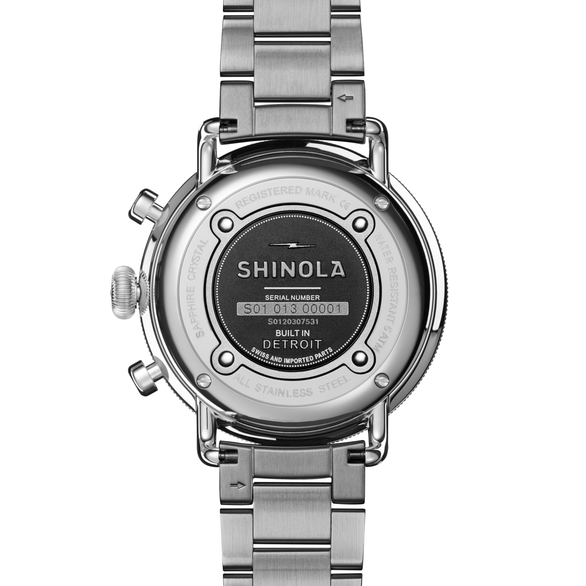 The back of the Shinola Canfield Chronograph Watch - Dark Olive displays the engraved brand name, serial number, and “Built in Detroit” text on its stainless steel case, along with the visible metal bracelet and clasp.
