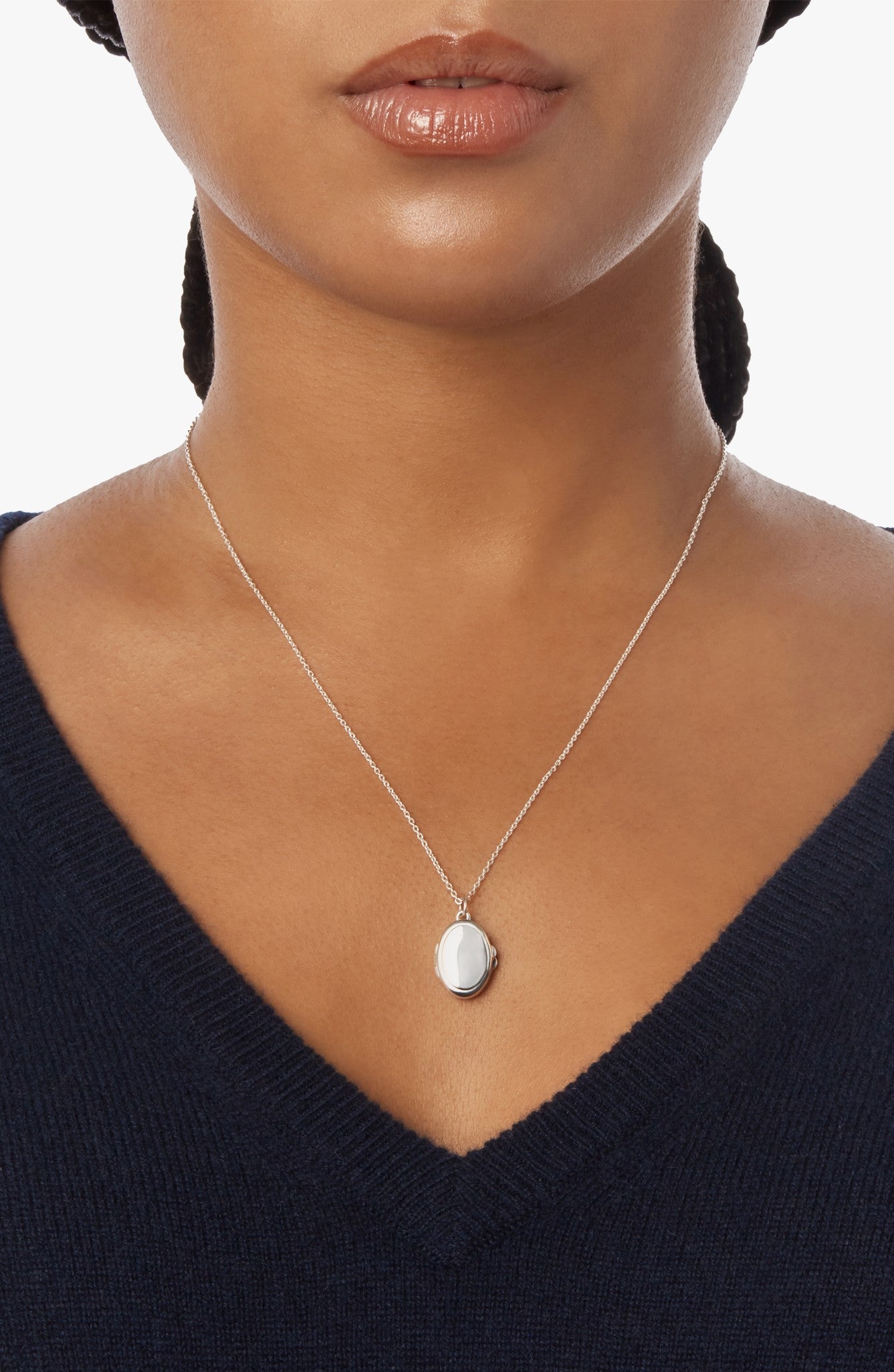 A woman wears the Shinola Heirloom Petite Locket Necklace in sterling silver, featuring an oval pendant, paired with a V-neck navy sweater. The photo highlights her neck, necklace, and lower face against a plain white background.