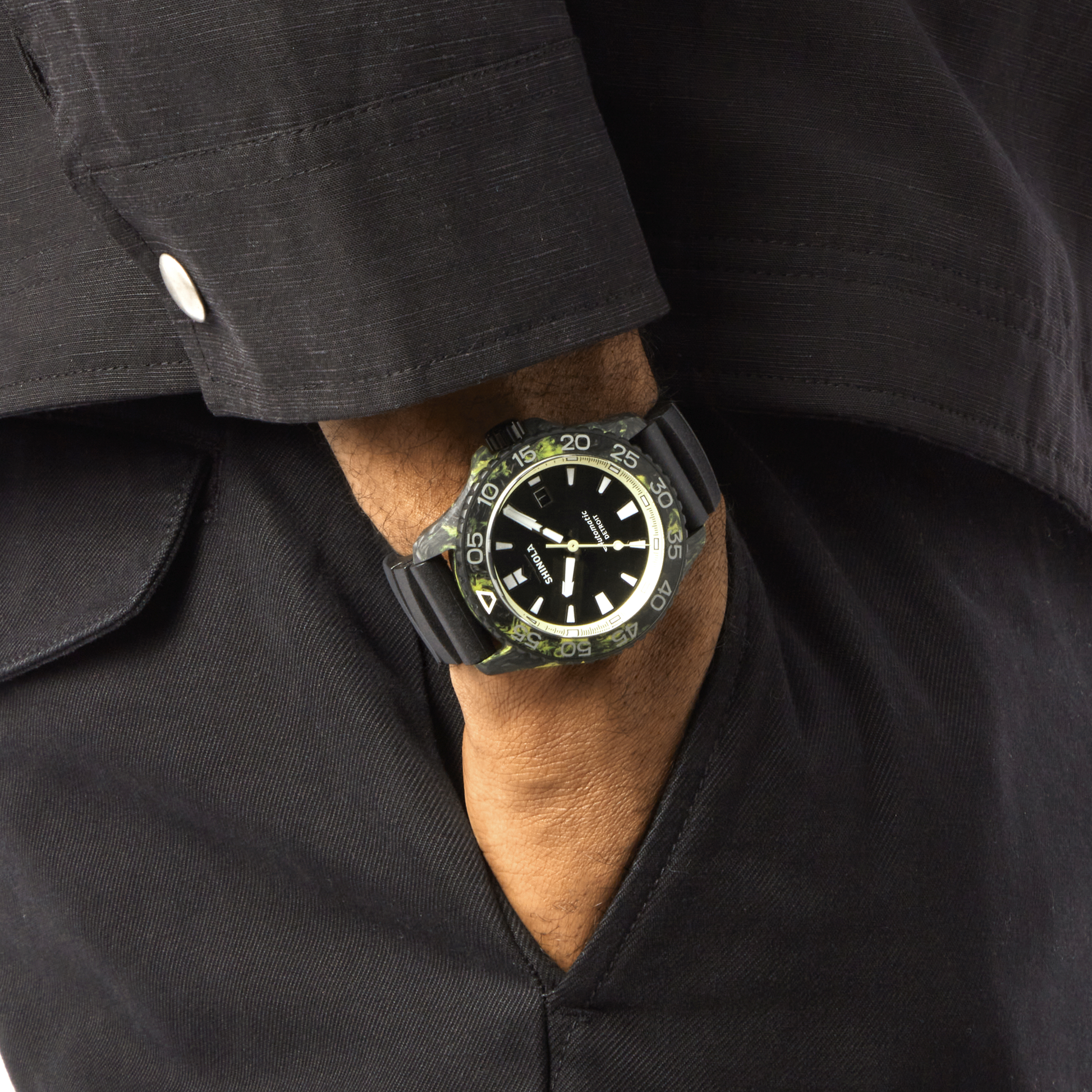A person in a black long-sleeve shirt and pants has their hand in their pocket, showing off the Shinola Forged Carbon Monster Watch with a round dial and luminous markers.
