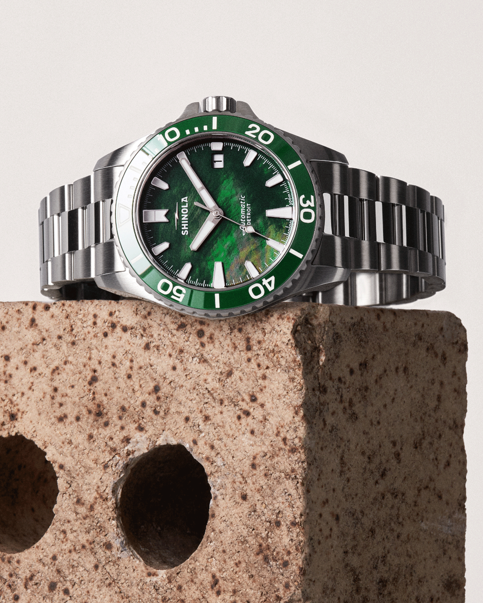 The Shinola Lake Ontario Monster Automatic Dive Watch, featuring a green bezel and dial, is displayed on a rough brown brick with two round holes against a plain light background.