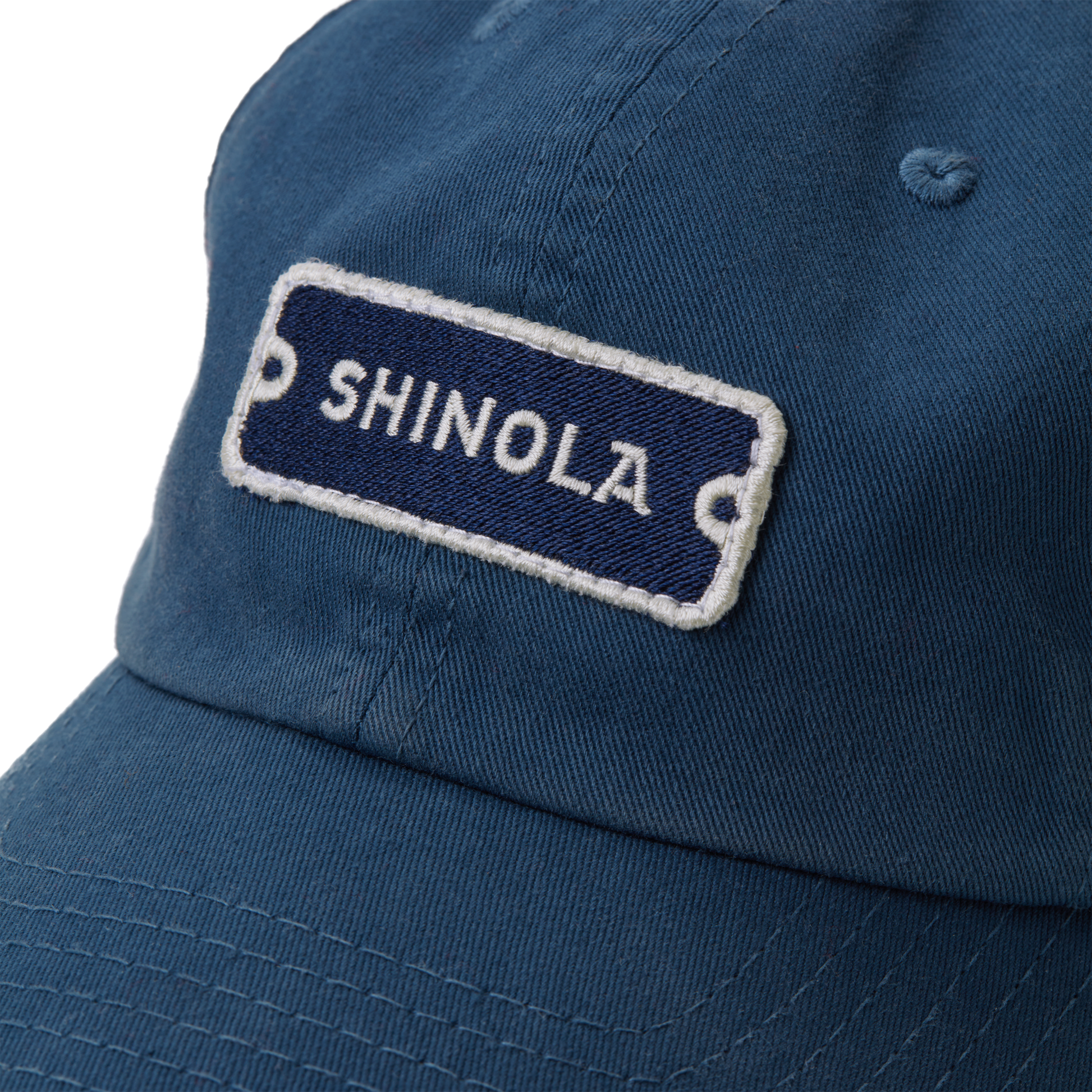 Close-up of the Shinola Cotton Twill 6 Panel Cap in blue, made from garment-dyed cotton twill, featuring a rectangular SHINOLA patch with white letters on a dark blue background and a leather strap closure.