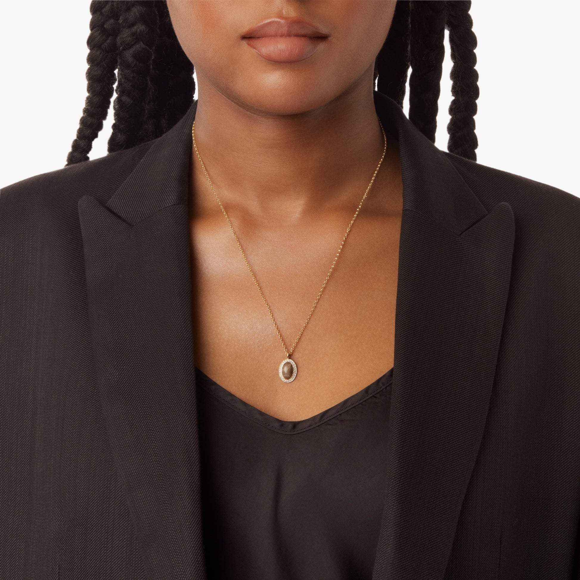 A woman with braided hair wears a black blazer and top, showcasing the Shinola Diamond Petoskey Petite Necklace in yellow gold. The necklace features an oval pendant, and the background is plain white.