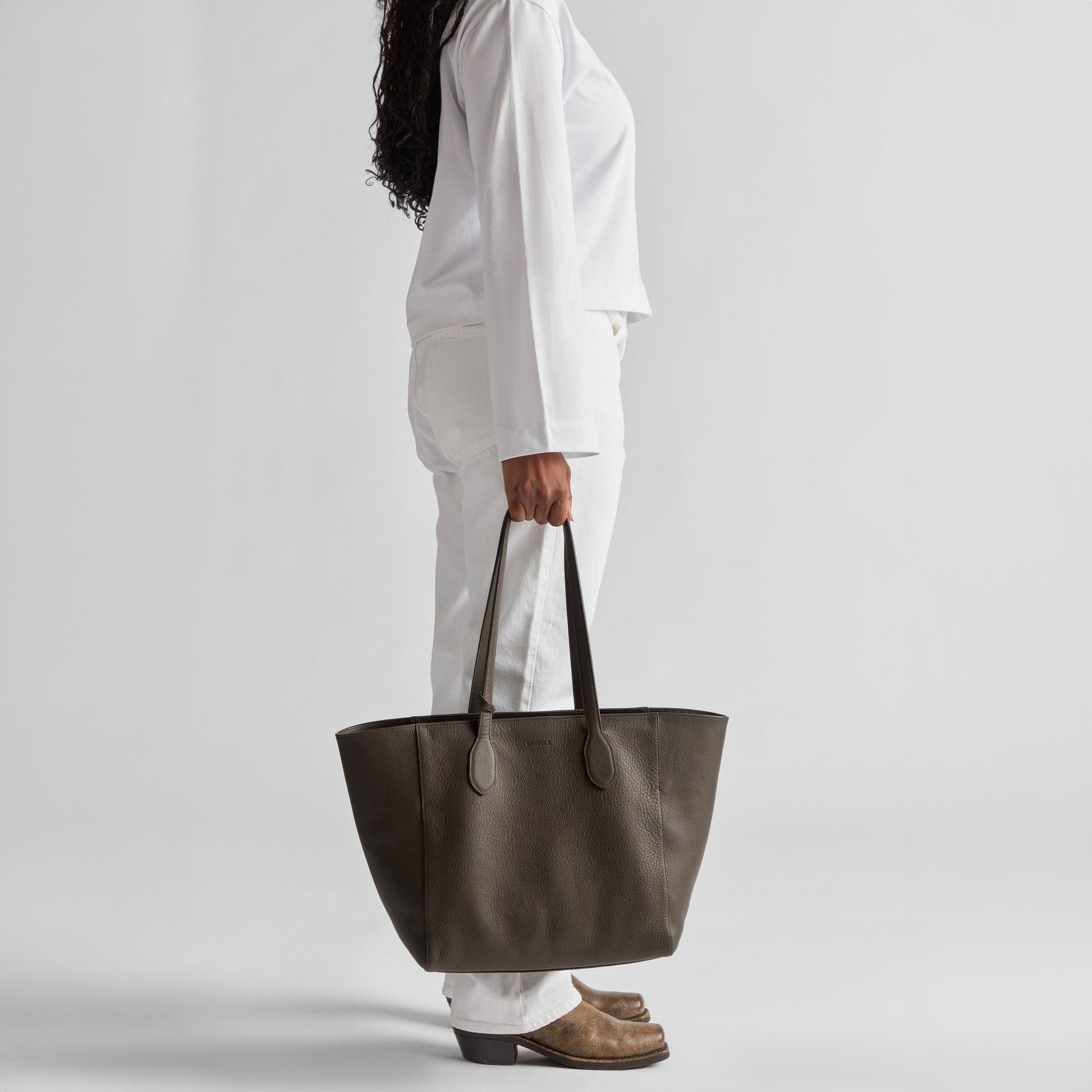 A person in white clothing and brown shoes stands in profile, holding the Shinola Runabout Tote Bag - Espresso by the handles, against a plain light background.