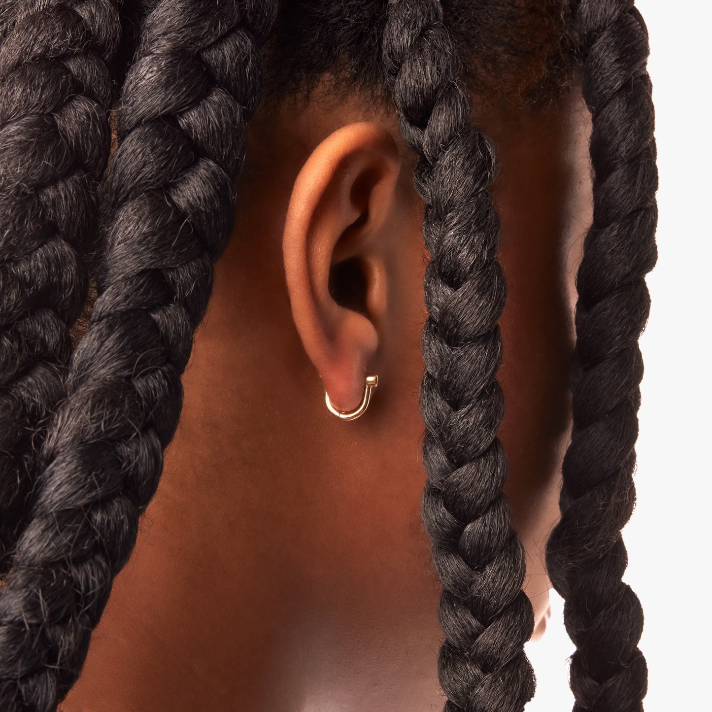 A close-up of a person’s ear wearing Shinola Bike Lock Huggie Earrings in yellow gold, framed by thick, dark braids against a light background.