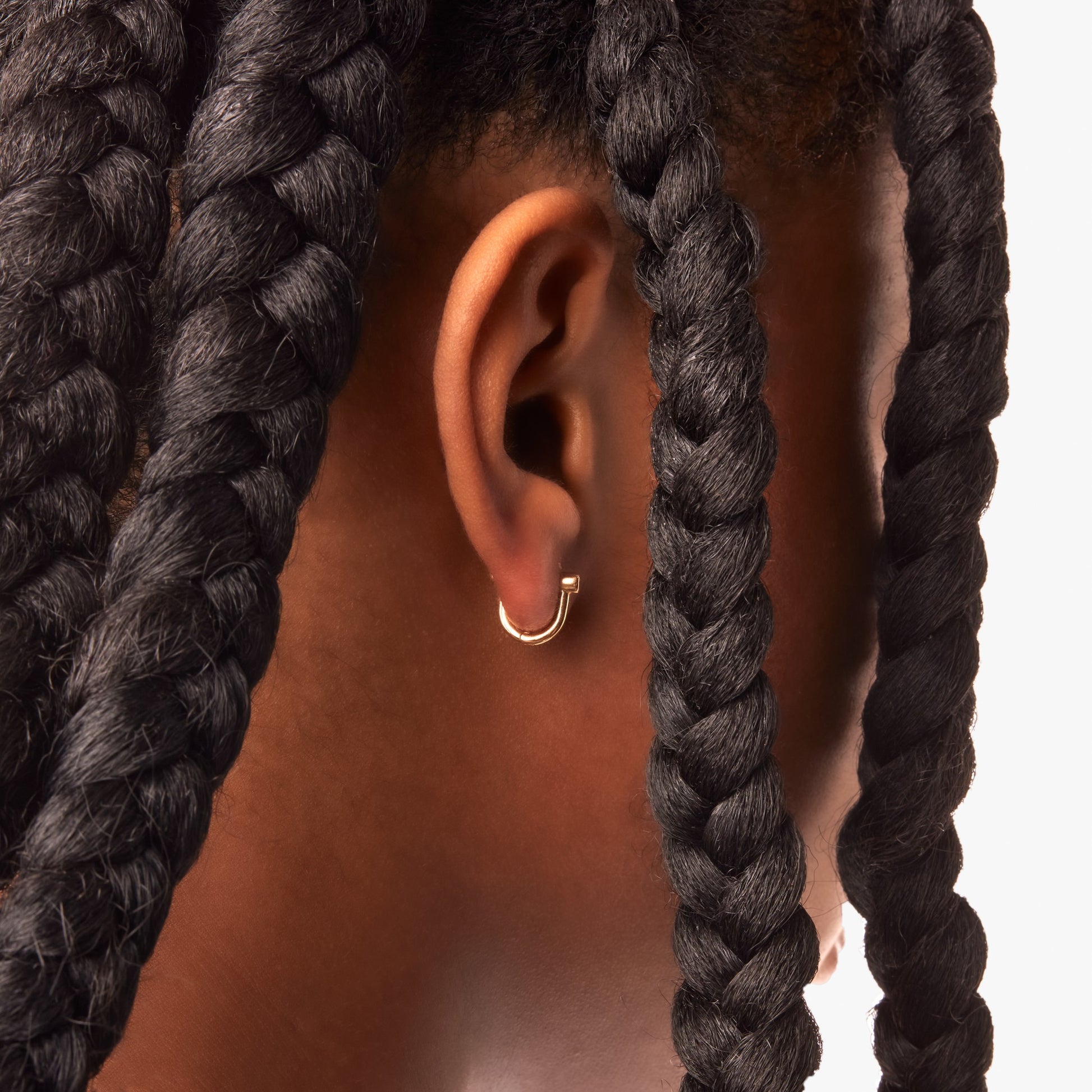 A close-up of a person’s ear wearing Shinola Bike Lock Huggie Earrings in yellow gold, framed by thick, dark braids against a light background.