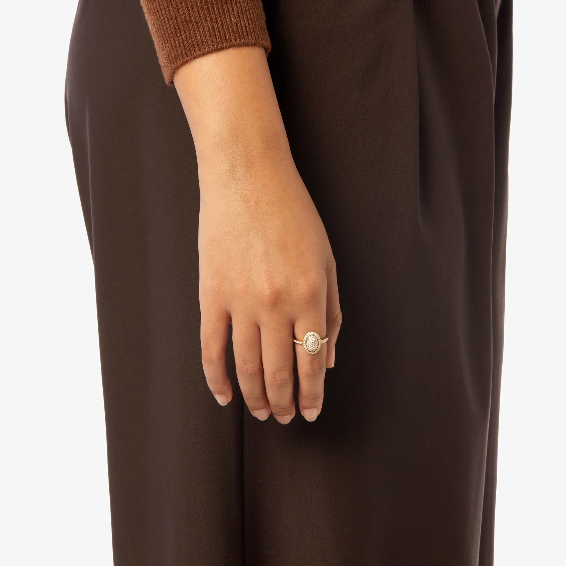 A close-up shows a hand wearing the Shinola Diamond Pave Oval Ring - Yellow Gold with natural diamonds, resting by their side. The person wears dark brown pants and a brown top against a plain background.