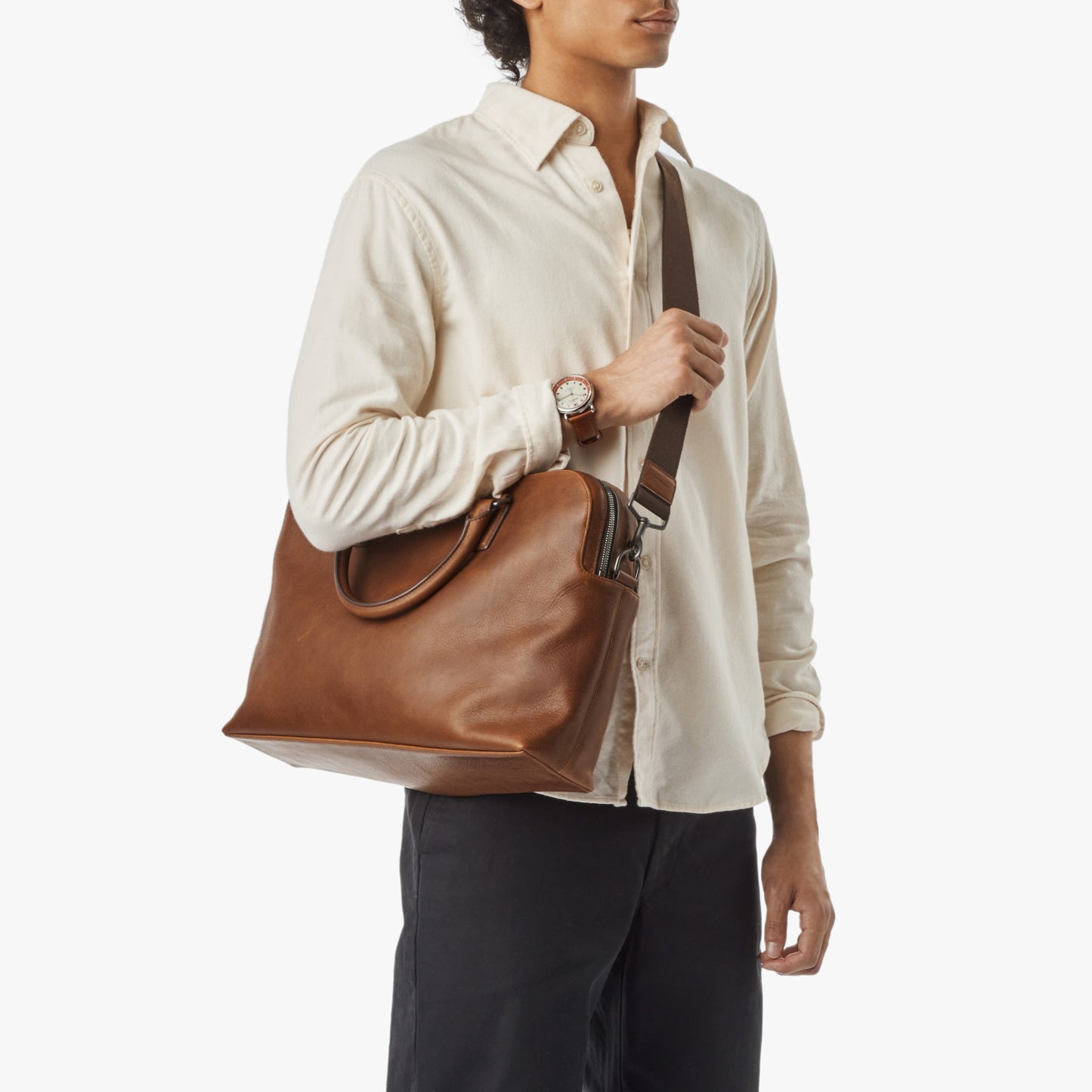 A person in a cream long-sleeve shirt and black pants carries the Shinola Double Zip Briefcase in Medium Brown by its handles and shoulder strap, perfectly matching their wristwatch against a plain white background.