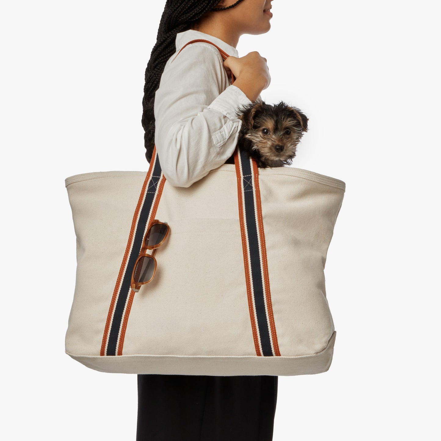 A woman carries a Shinola Large Easy Canvas Tote - Natural, with striped handles, sunglasses hooked on the front, and a small puppy peeking out from its spacious open compartment.