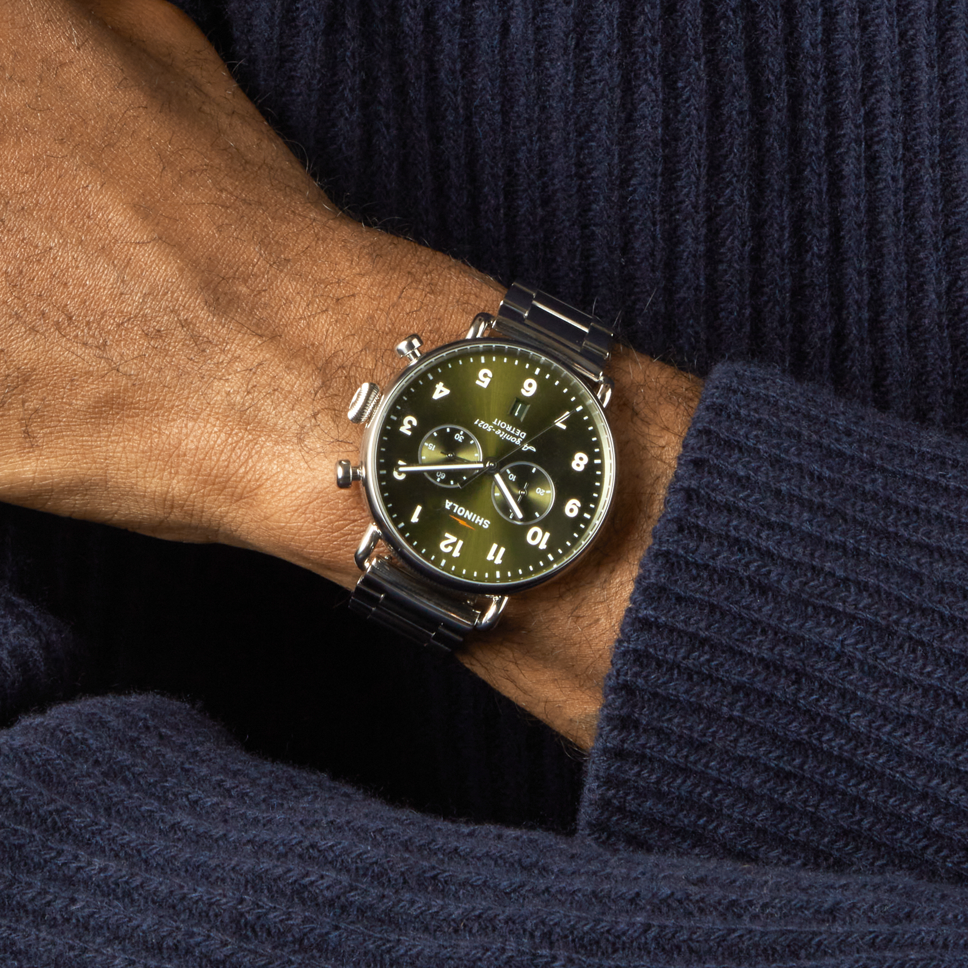 A person in a dark blue sweater wears a Shinola Canfield Chronograph Watch - Dark Olive, featuring a stainless steel case, black strap, white Arabic numerals, and two subdials on the left wrist atop the right arm.