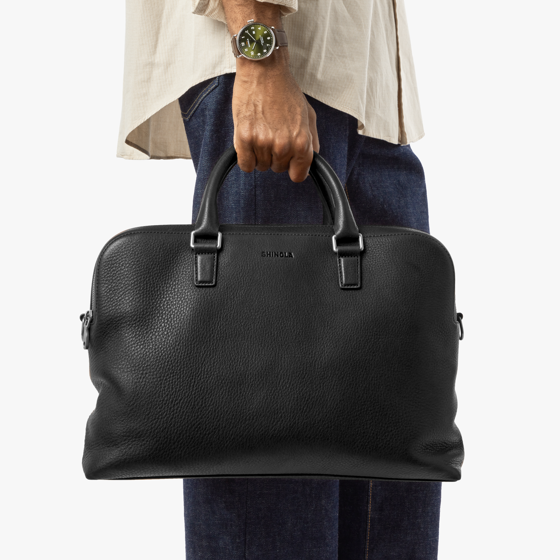 A person in a beige shirt, blue jeans, and wristwatch holds a Shinola Double Zip Briefcase - Black by the handles, showing its extra compartment against a plain background.