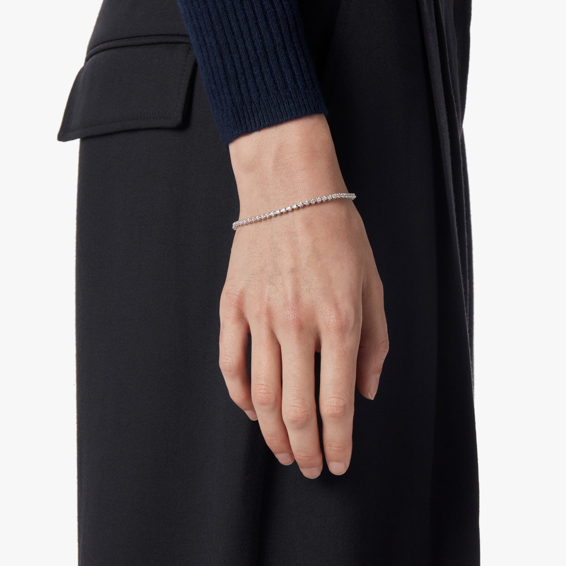 A close-up shows a hand and wrist wearing the Shinola 1 Carat 7" Diamond Tennis Bracelet in white gold. The person is dressed in a dark long-sleeve top and pants, set against a plain white background.