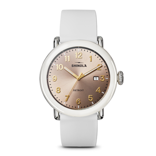 The Shinola Detrola Watch - Nude features a round, rose-gold dial with gold numerals and hands, a small date window, and a white leather strap, embodying classic American design.