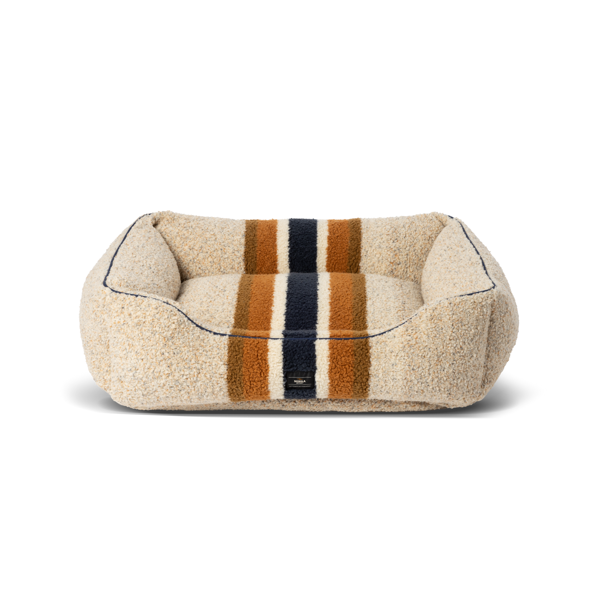 The Shinola Kuddle Dog Bed - Stripe Oat is a rectangular, plush beige bed with orange, navy blue, and brown stripes, raised sides, soft textured surface, memory fiber fill, minimalist design, and a washable cover.