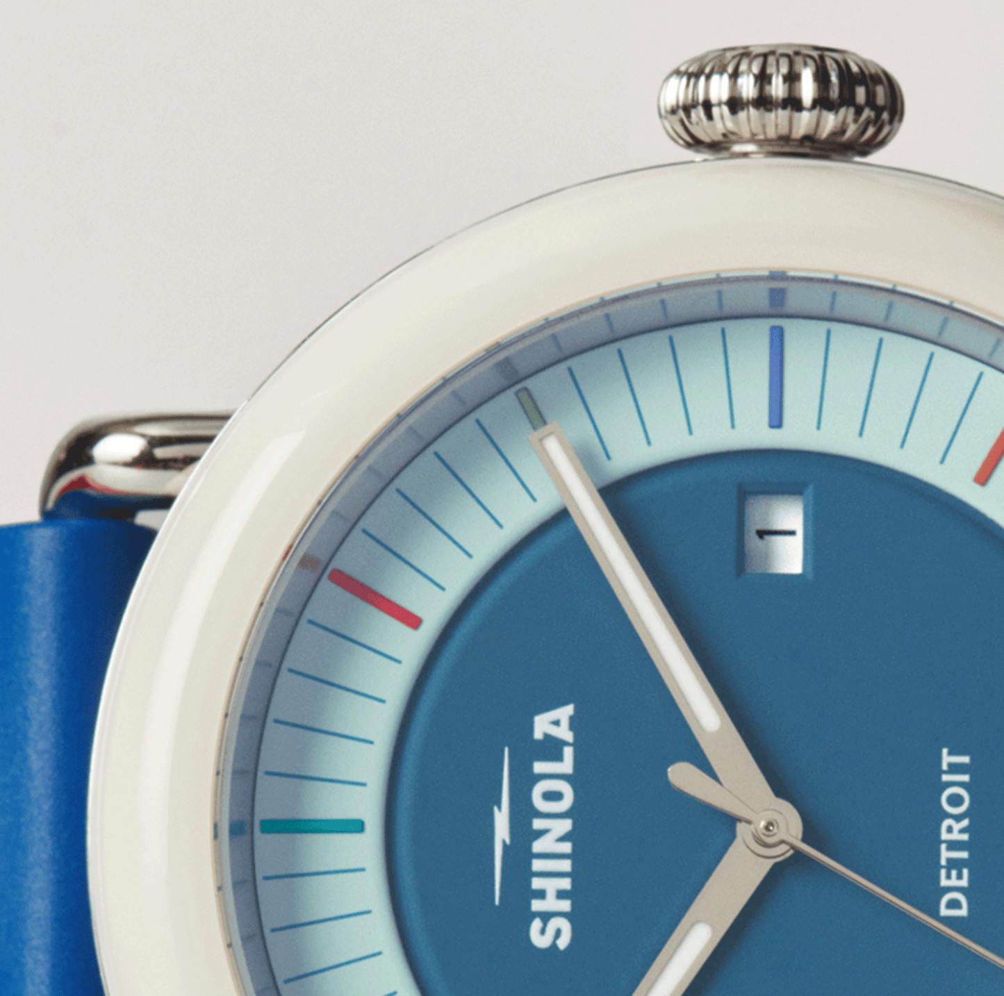 Close-up of the Shinola Detrola Watch - Blue, featuring a white bezel, colorful hour markers, small date window showing “1,” and a blue strap. The watch face displays 1:05.