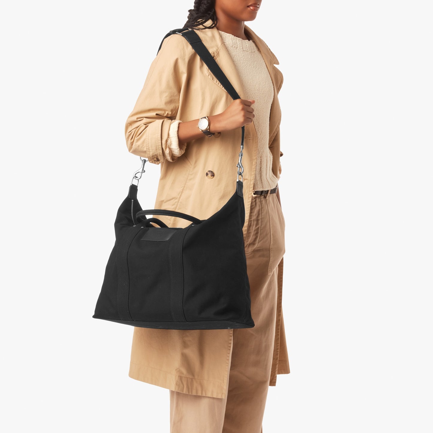 A person in a beige trench coat and tan pants carries the Shinola Runabout Holdall Canvas Bag - Black, featuring short handles and a crossbody strap, against a plain white background.
