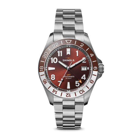 The Shinola Monster GMT Automatic Watch - Whiskey features a stainless steel case, metallic red-brown dial, silver and white hour markers, date at 3 o’clock, 24-hour GMT bezel, and a silver link bracelet.