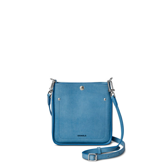 The Shinola Mini Pocket Crossbody Bag - River is a small, rectangular, light blue bag with a front pocket, silver hardware, and adjustable strap, shown against a plain white background.