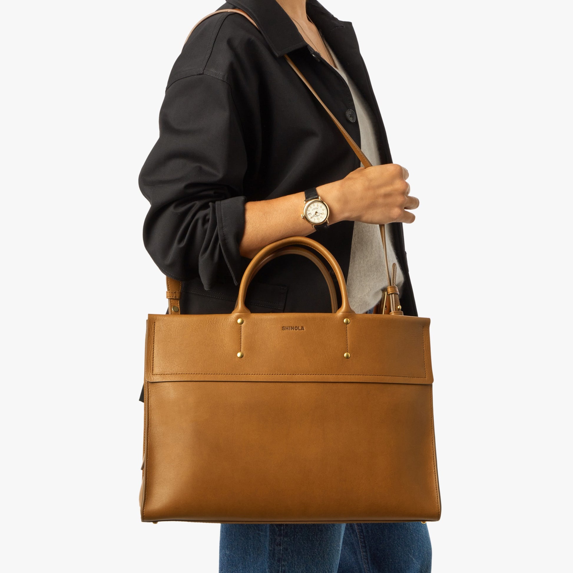 A person in a black jacket, gray shirt, and blue jeans carries the Shinola Day Satchel - Tan, a spacious leather bag with top handles, shoulder strap, gold accents, laptop compartment, and "Shinola" on the front.