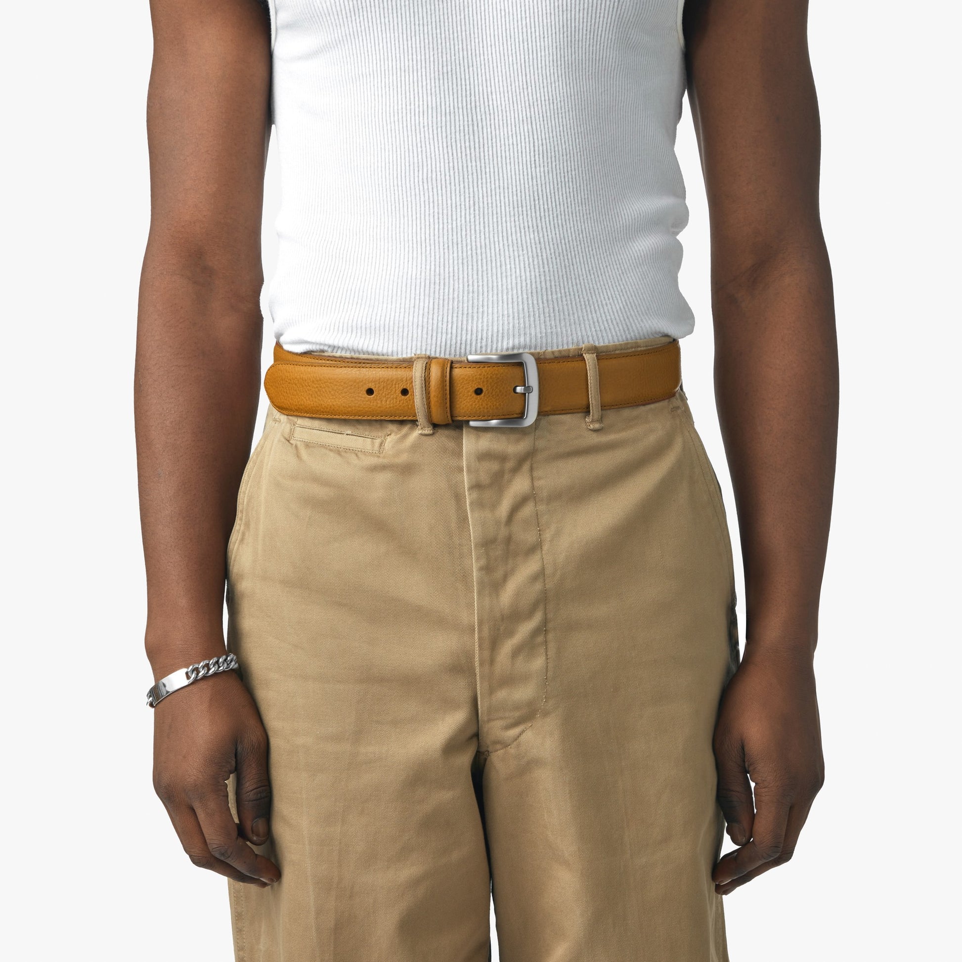 A person wearing a white ribbed tank top, tan high-waisted pants, and the Shinola 1 1/2" Canfield Belt in Bourbon with a silver buckle, accessorized with a chain bracelet, stands against a plain white background.