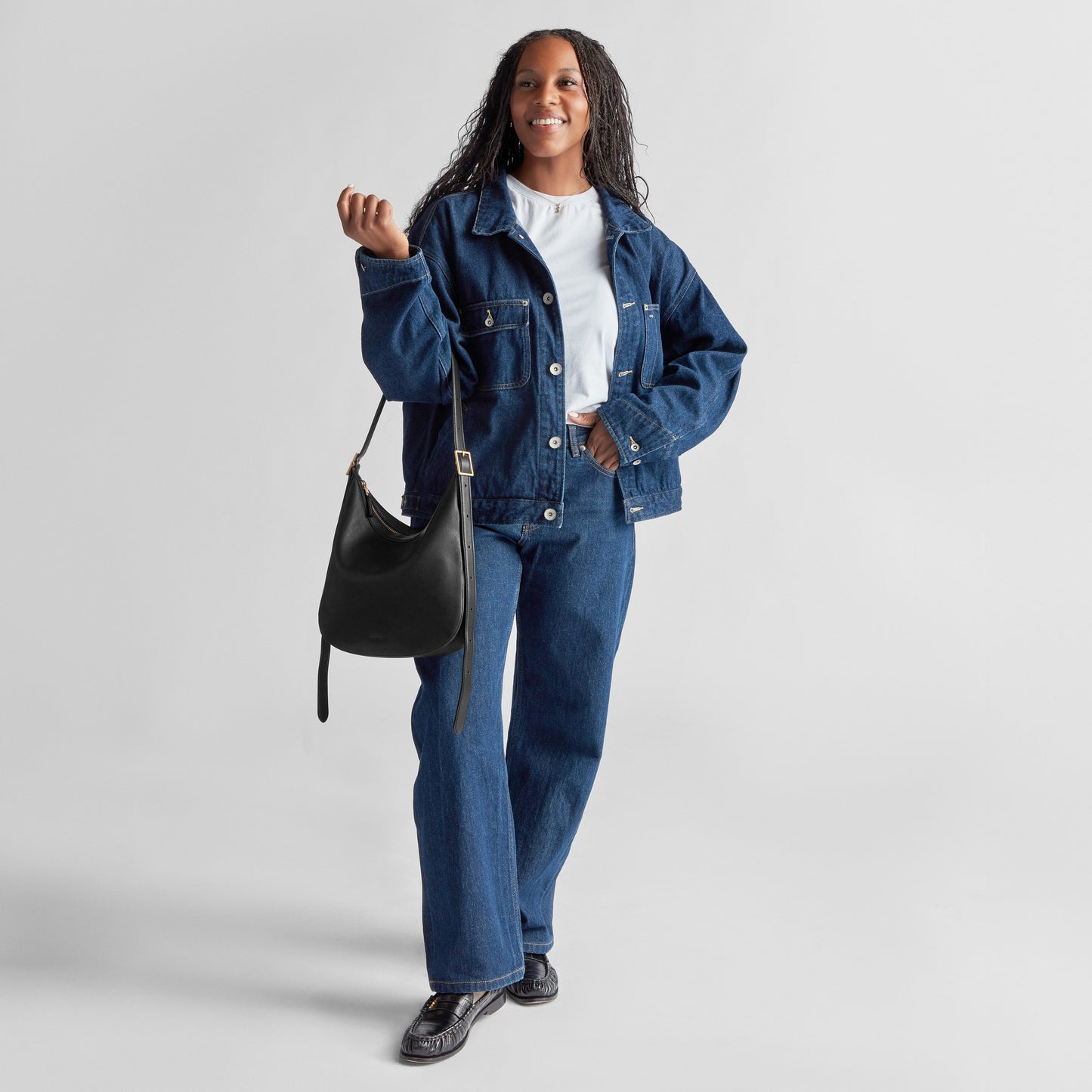 A woman in a blue denim jacket, jeans, white top, and black shoes smiles while holding the Shinola Medium Bixby Crossbody - Black—a versatile natural leather accessory—against a plain light background.