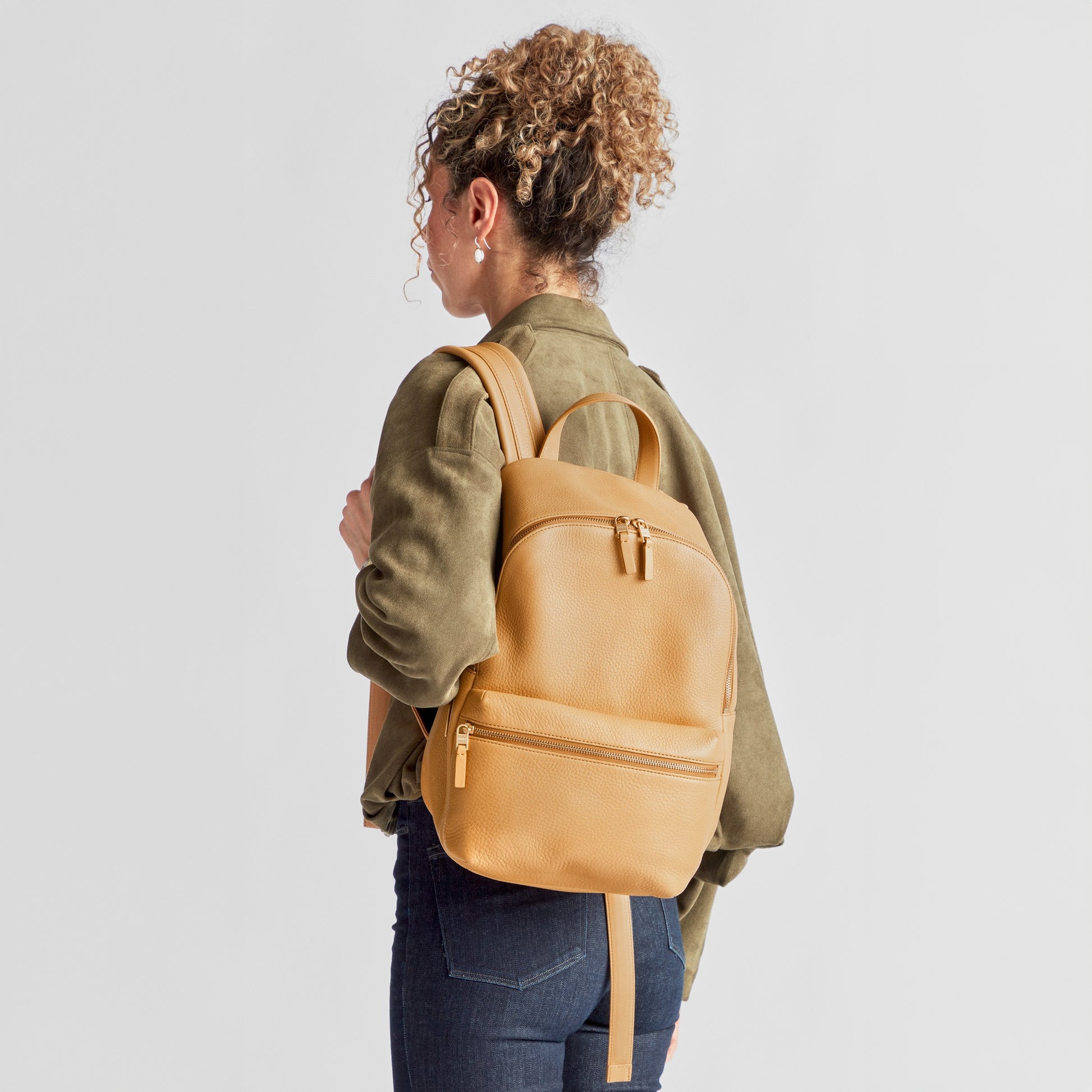 A person with curly hair, in a green jacket and jeans, is seen from behind carrying the Shinola Small Runwell Backpack - Honey, crafted from Natural Grain Leather, against a plain light background.