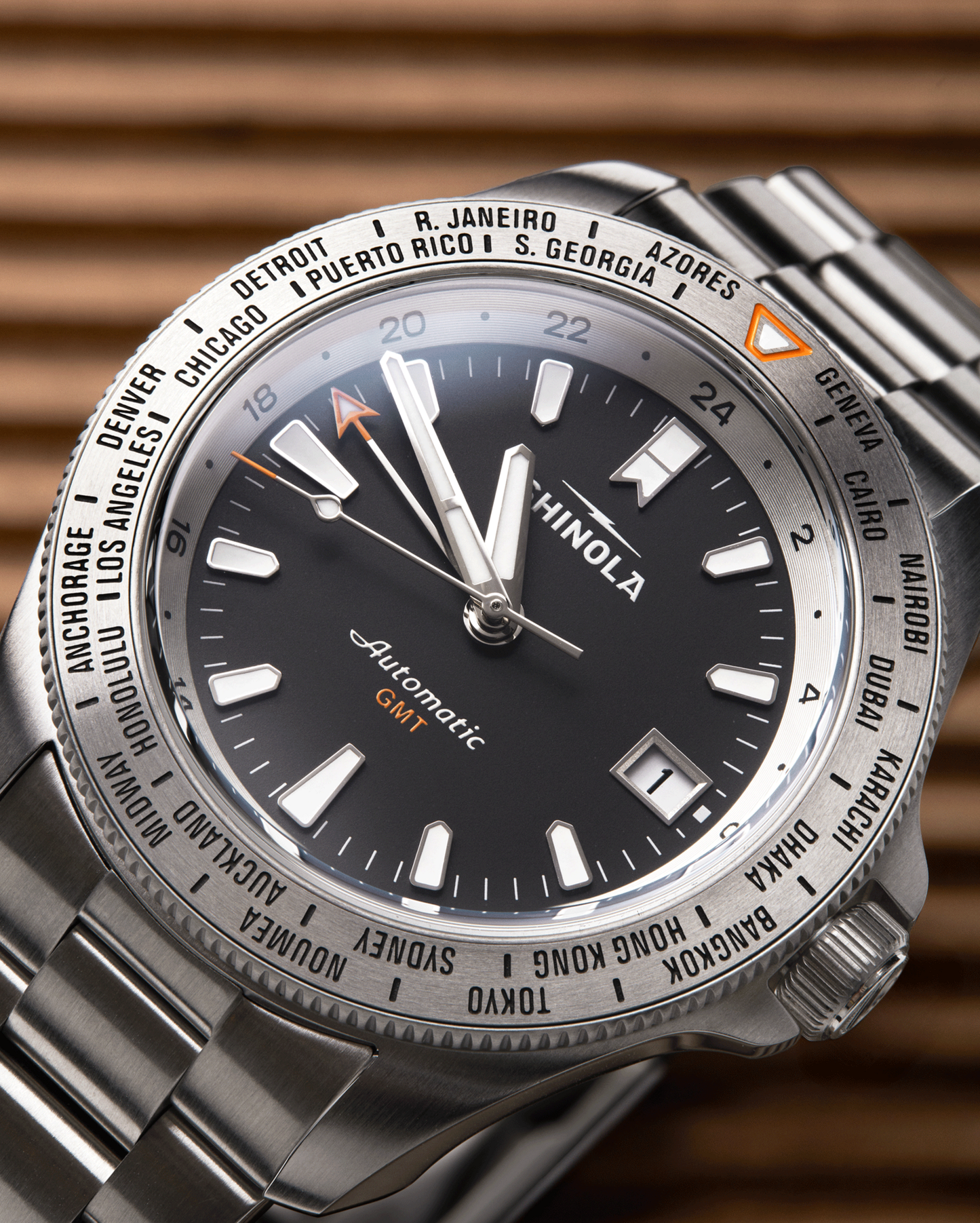 A close-up of a Shinola Monster Worldtimer GMT Watch with a stainless steel band, black dial, silver hour markers, date window, orange GMT hand, and world cities engraved on the bezel.