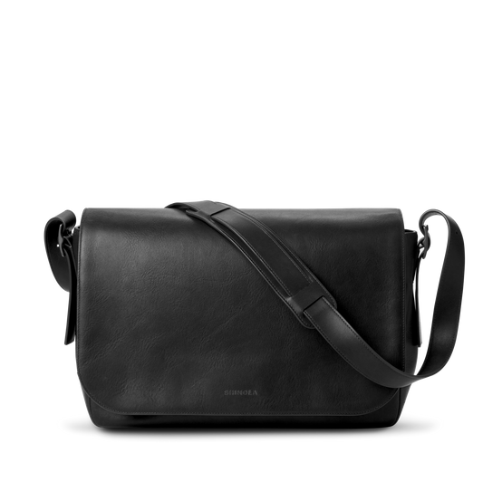 The Shinola Runwell Messenger Bag in black features a flap closure, padded laptop sleeve, and adjustable crossbody strap, displayed against a plain white background.