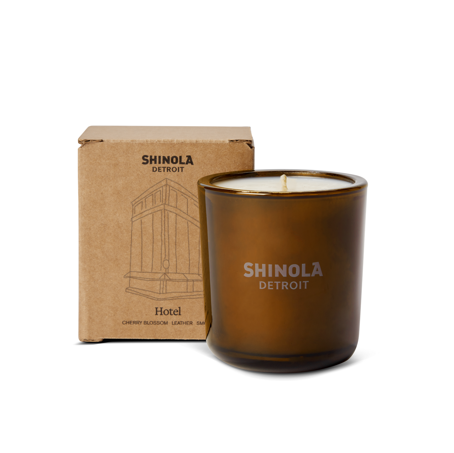 A brown recycled glass Shinola "Hotel" 8oz scented candle sits before a matching box with a cabinet design. Crafted from coconut soy wax, it features cherry blossom, leather, and sage notes.