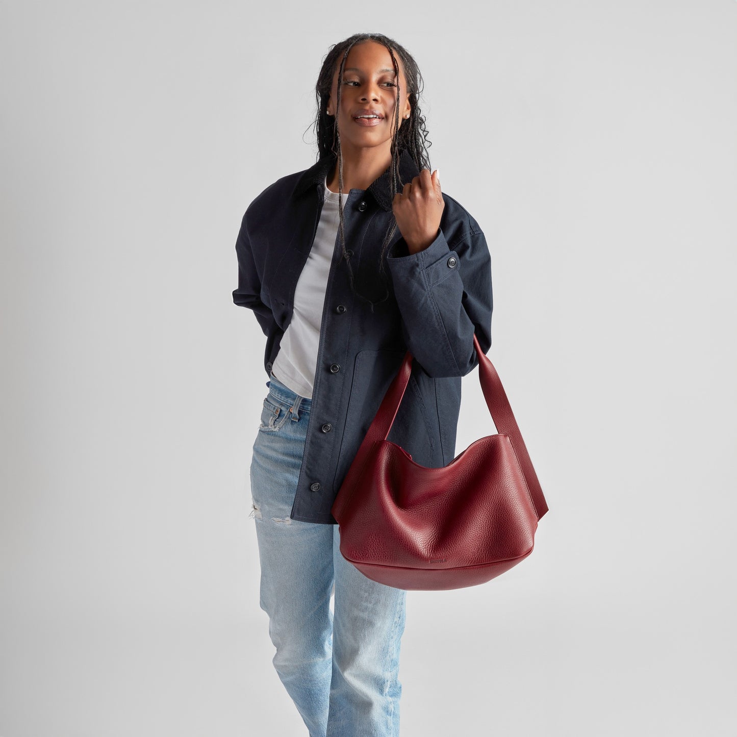 A person with long braids, in a navy jacket, white shirt, and ripped light blue jeans, smiles while holding the Shinola Medium Derby Shoulder Bag in Ruby. The background is plain light gray.