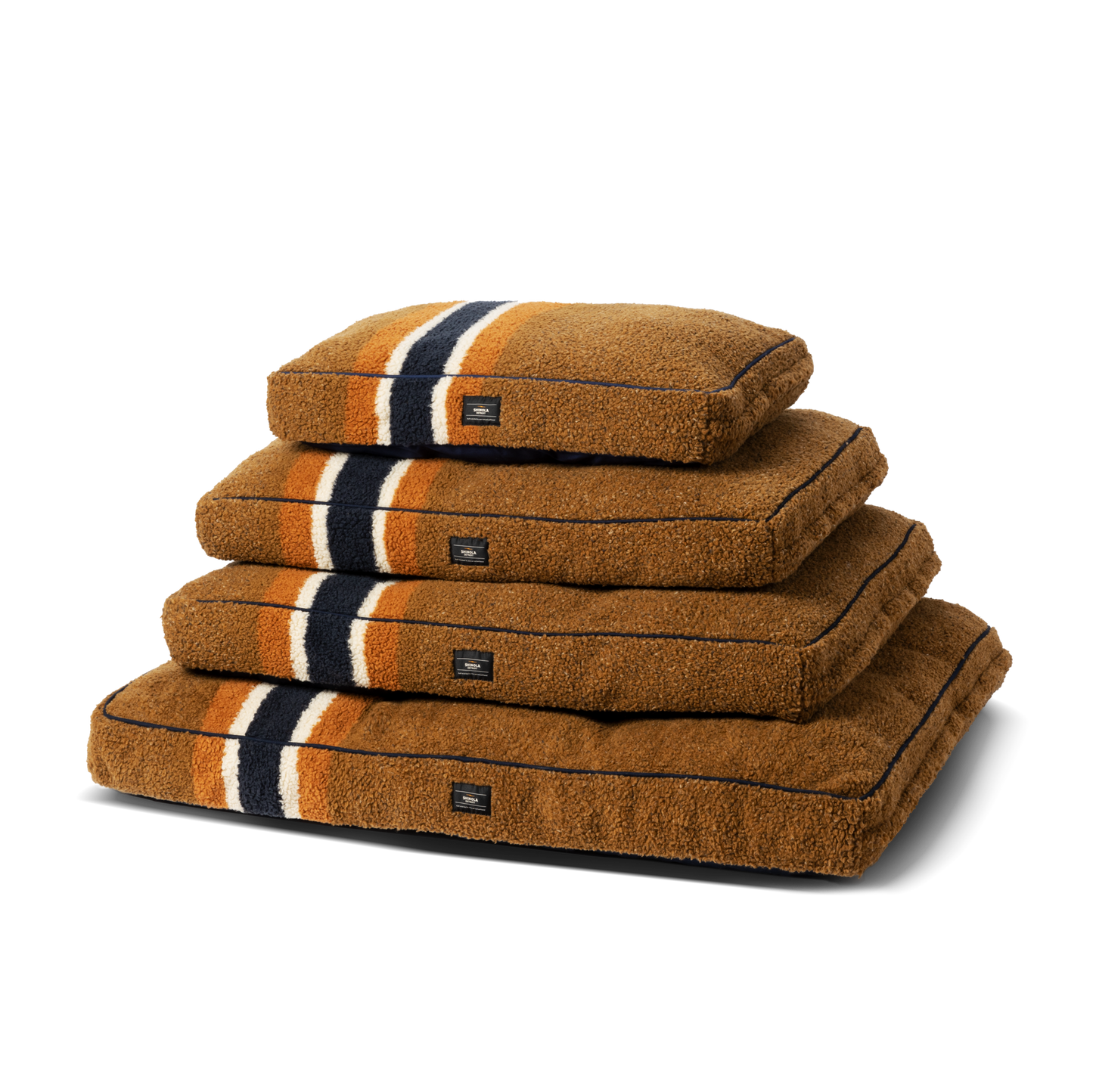 Four Shinola Napper Pillow Dog Beds in stripe brown are stacked, each with black, white, and orange stripes and a black front label. These beds feature memory fiber fill and have machine-washable covers for easy care.