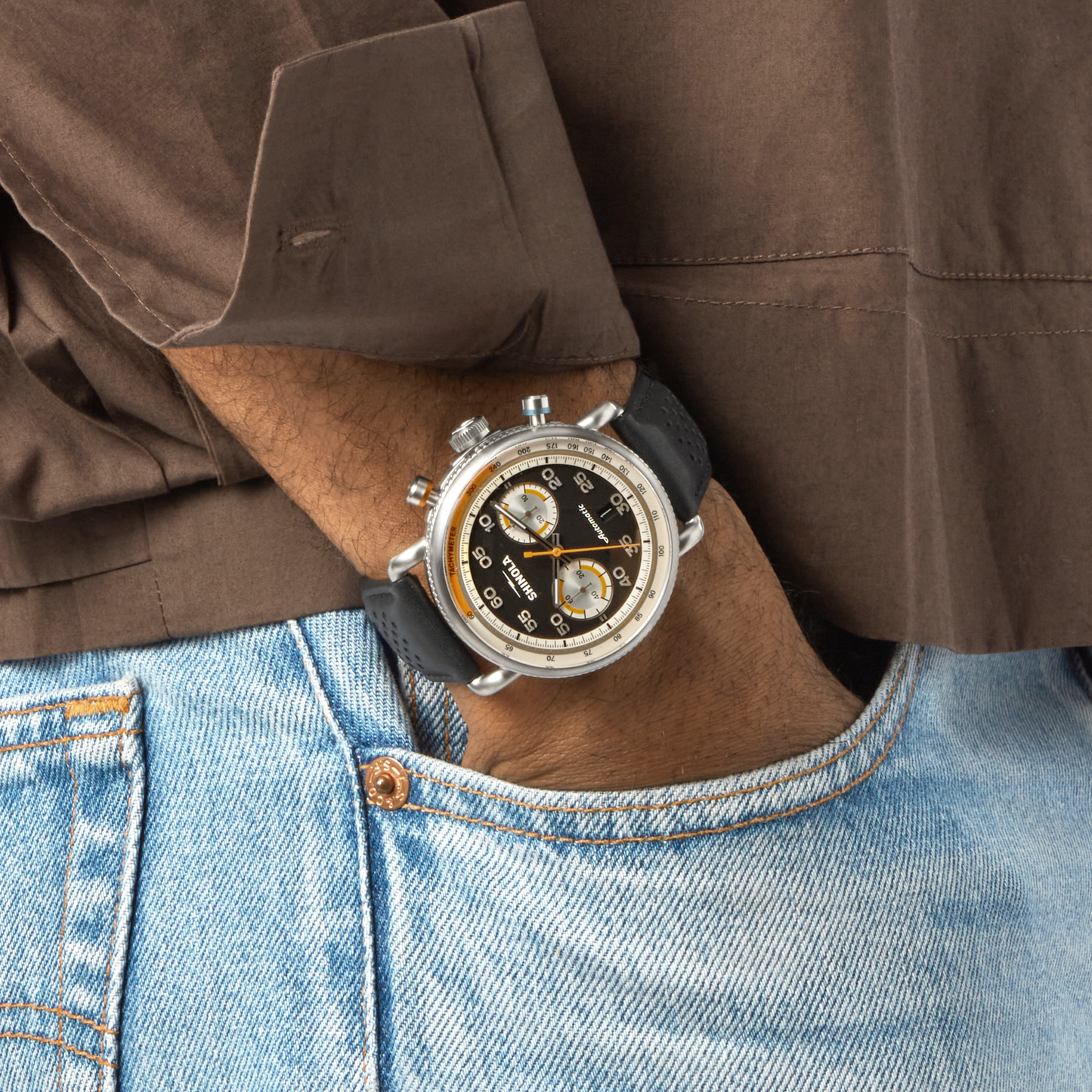 A person in a brown shirt and blue jeans rests their hand in their pocket, showcasing the Shinola Canfield Speedway Open Road Automatic Chronograph Watch in black, featuring multiple dials, a black strap, and Swiss-made automatic movement.