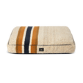 The Shinola Napper Pillow Dog Bed - Stripe Oat is a rectangular beige bed with a fuzzy Berber cover, dark blue, gold, and brown horizontal stripes, a small black front label, and is machine washable. Shown on a plain white background.