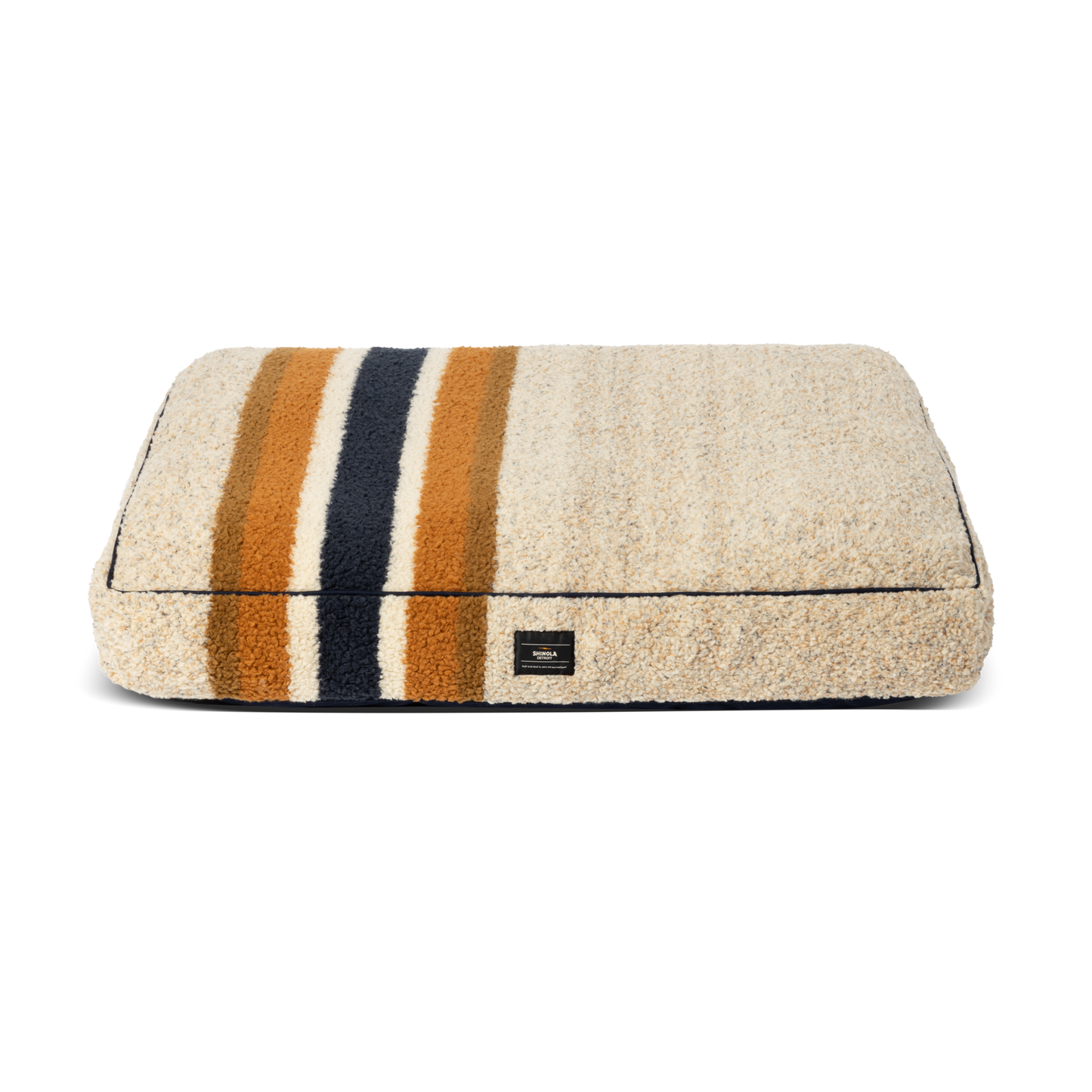 The Shinola Napper Pillow Dog Bed - Stripe Oat is a rectangular beige bed with a fuzzy Berber cover, dark blue, gold, and brown horizontal stripes, a small black front label, and is machine washable. Shown on a plain white background.