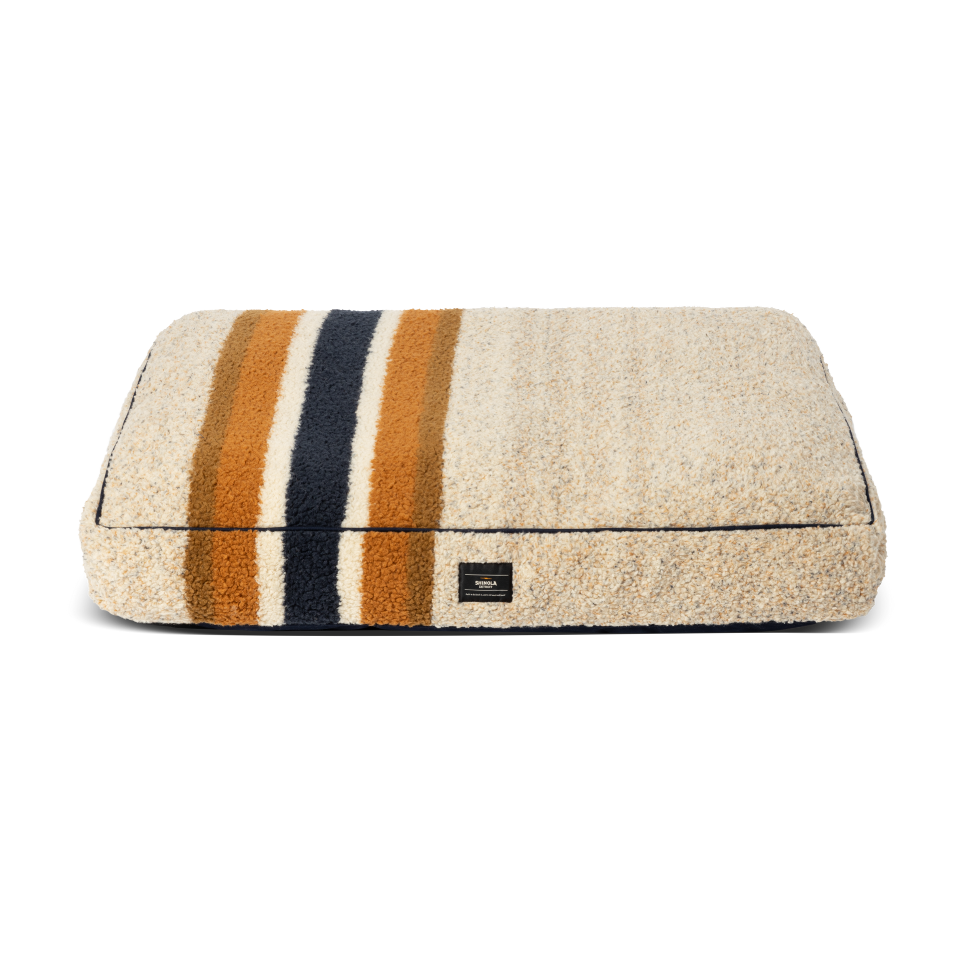 The Shinola Napper Pillow Dog Bed - Stripe Oat is a rectangular beige bed with a fuzzy Berber cover, dark blue, gold, and brown horizontal stripes, a small black front label, and is machine washable. Shown on a plain white background.