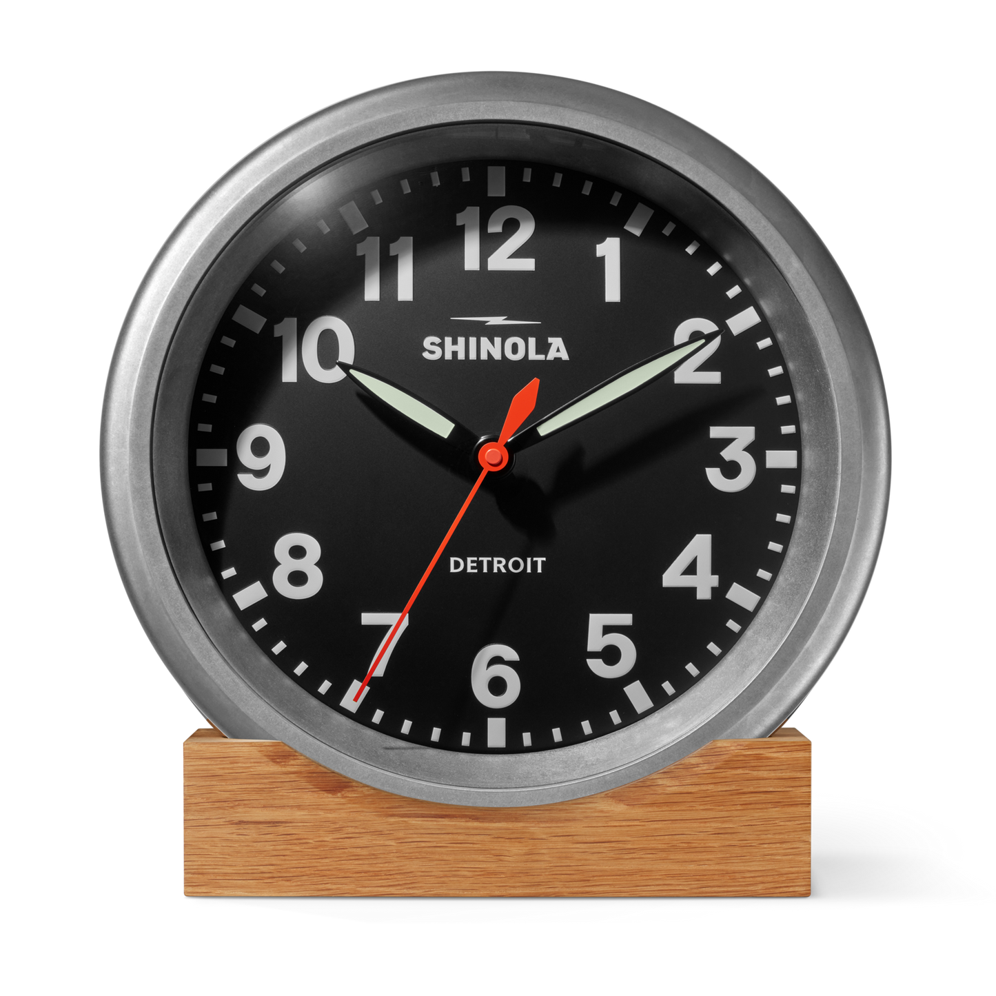 A Shinola 14" Runwell Clock in tumbled zinc with a black face, white numbers, and silver frame sits on a wooden base, inspired by industrial style and displaying 10:10:33.
