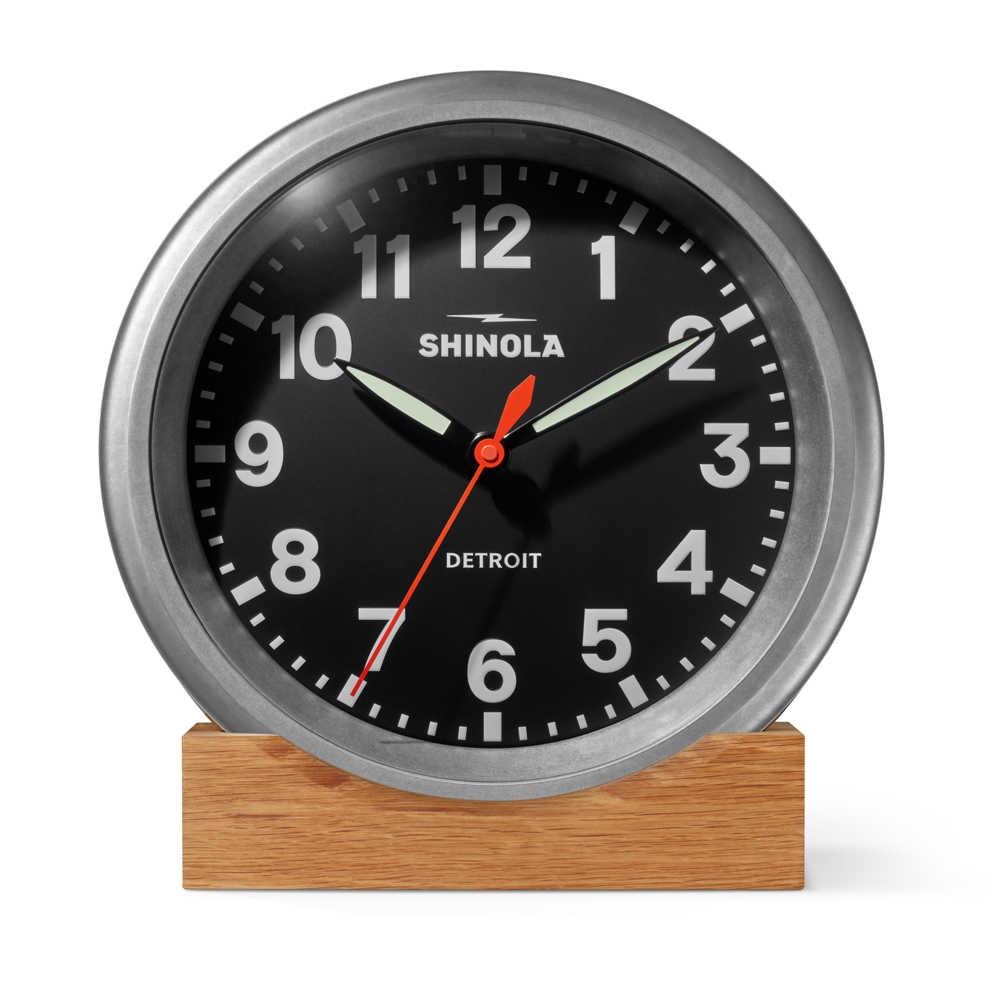 A Shinola 14" Runwell Clock in tumbled zinc with a black face, white numbers, and silver frame sits on a wooden base, inspired by industrial style and displaying 10:10:33.