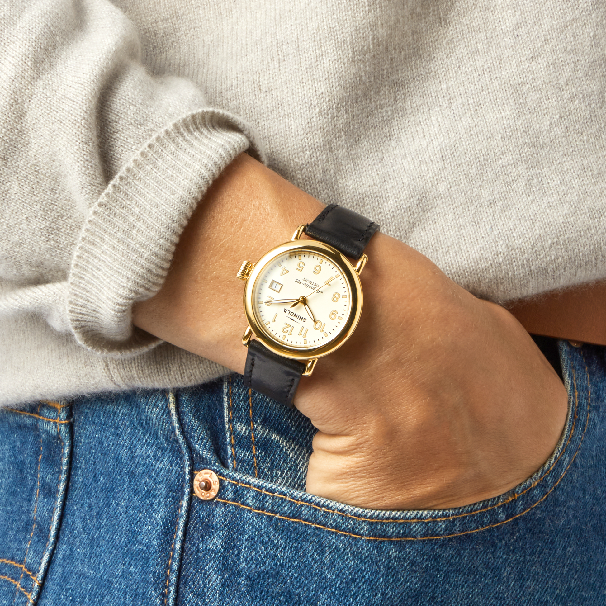 A person wearing the Shinola Runwell Watch - Alabaster with a gold stainless steel case, black leather strap, and cream dial, dressed in a light gray sweater and blue jeans, casually rests their hand in a jeans pocket.